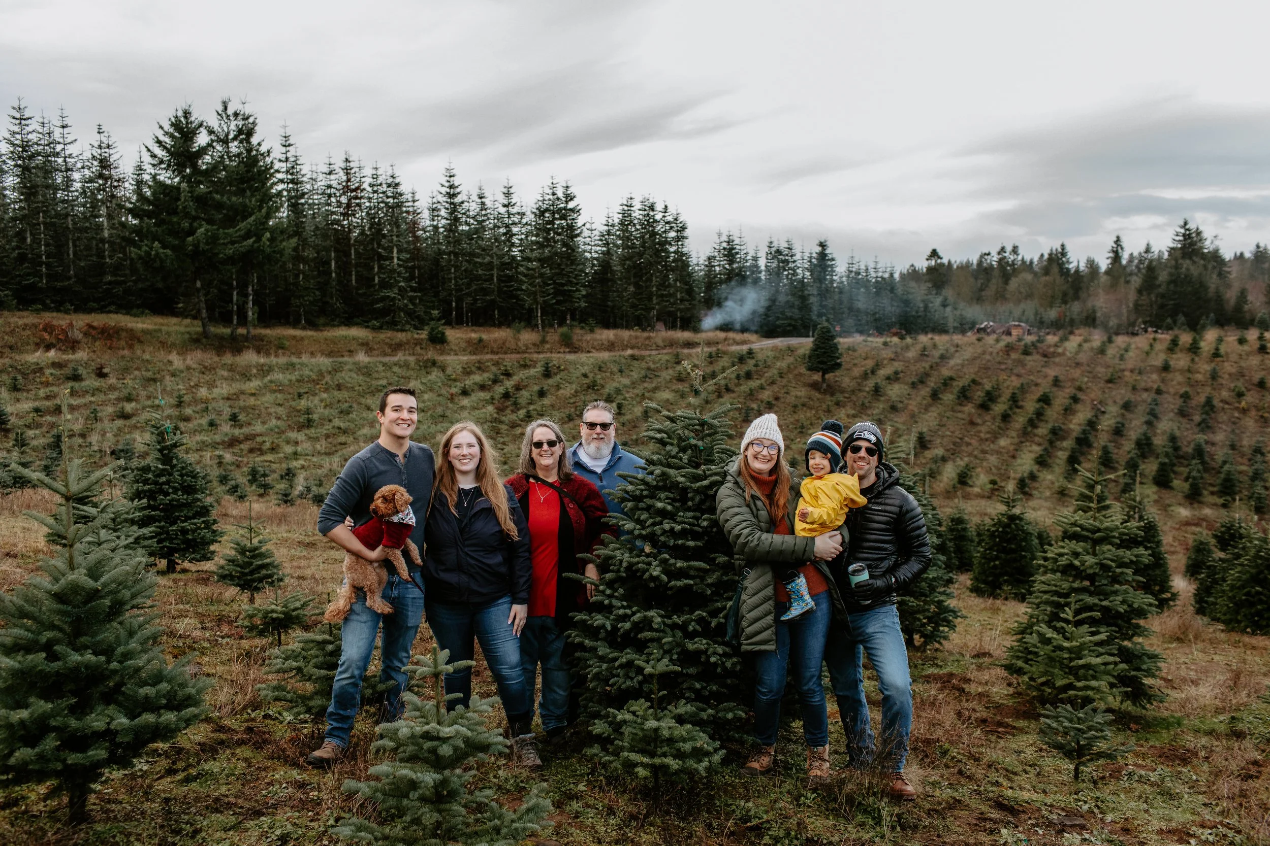 Family and friends at a Christmas tree farm, standing next to a pine tree, with a backdrop of sloping hills and a forest.