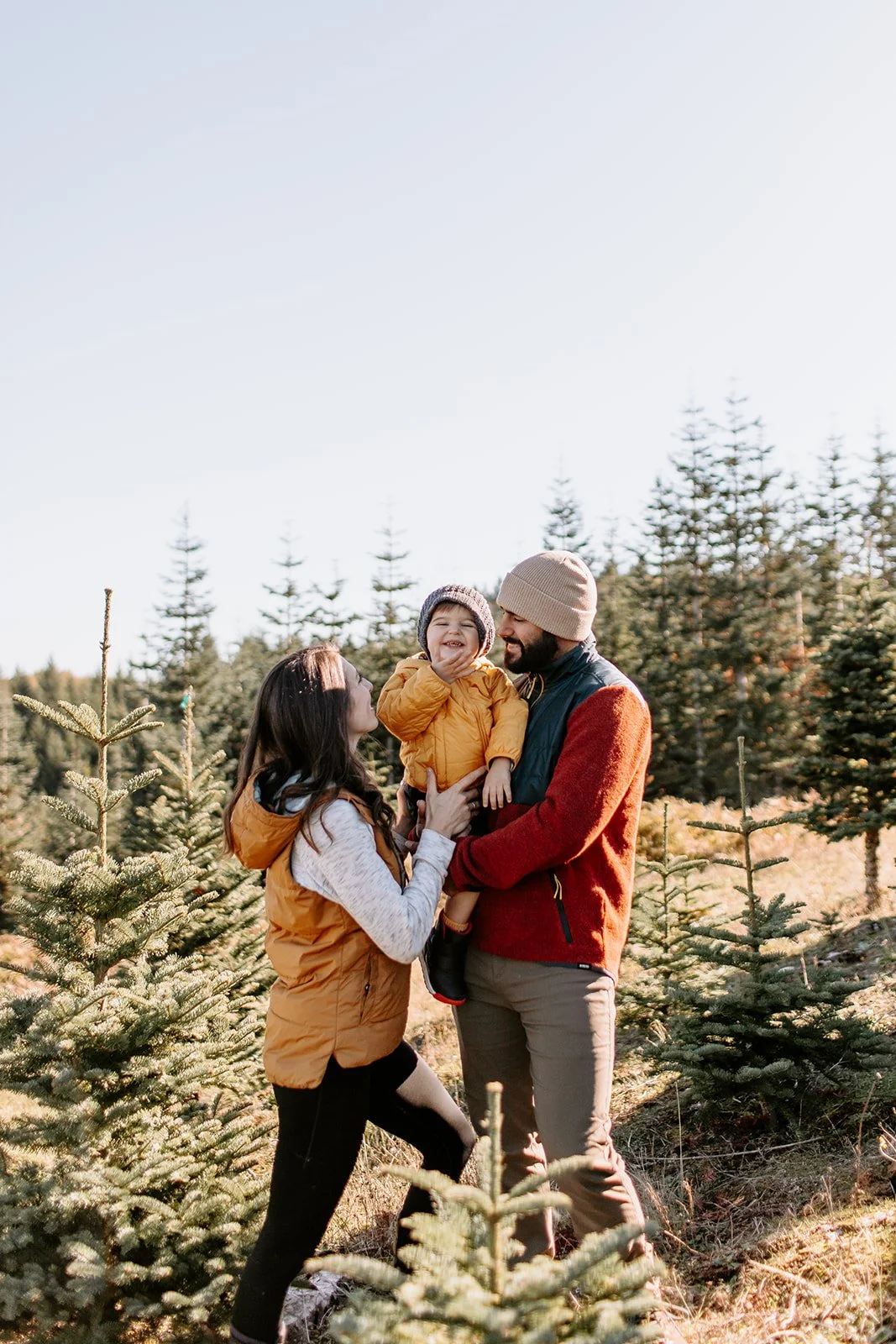 A family of three enjoying time outdoors among Christmas trees, with a woman and man holding a smiling young child between them.