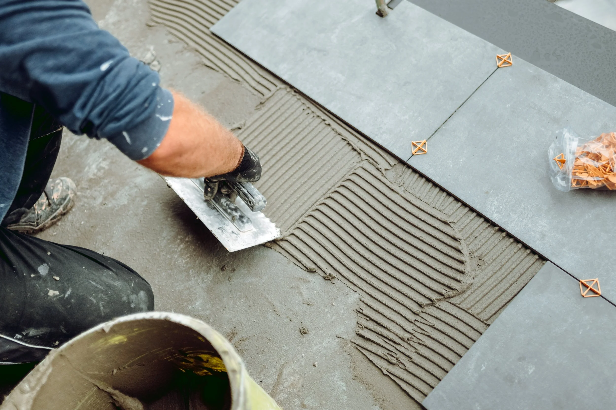 A construction worker applies tile adhesive with a trowel on a concrete surface, preparing to lay floor tiles. Construction tools and other tiles are visible.