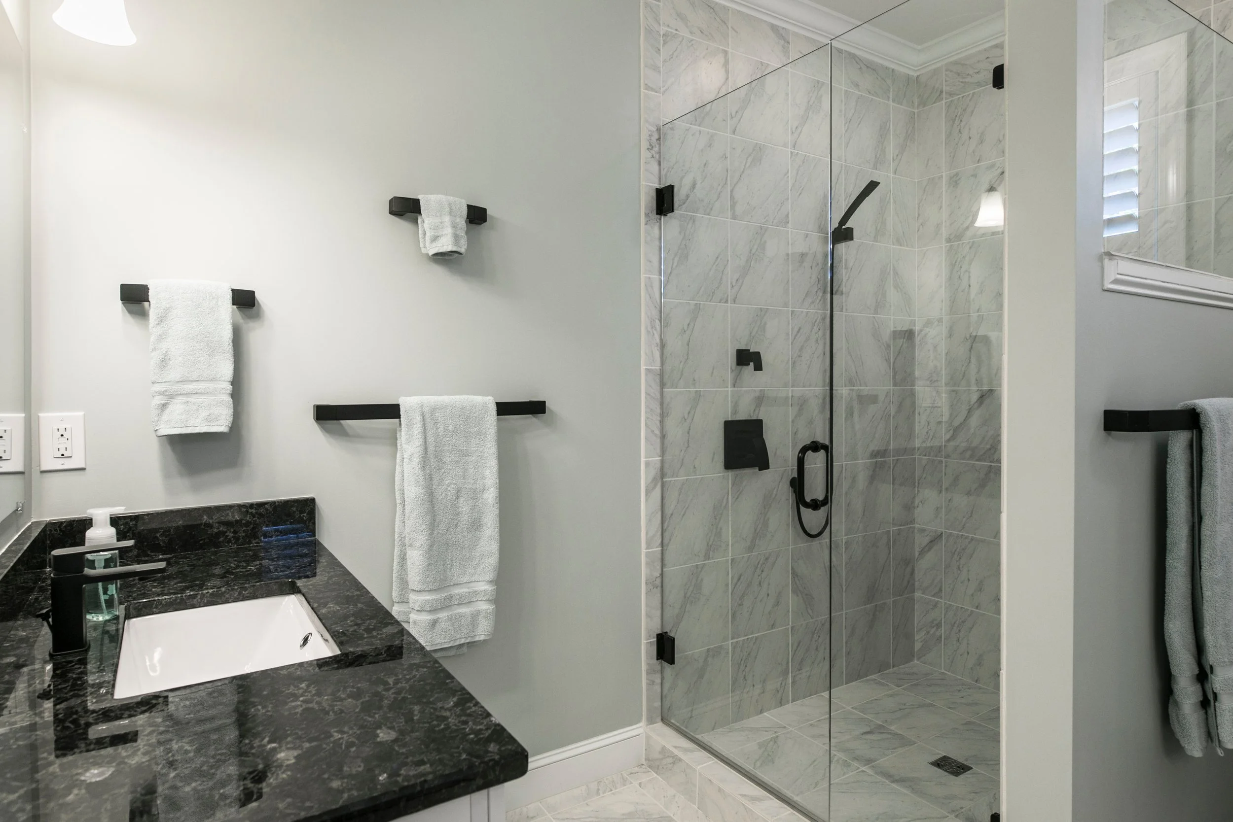 Modern bathroom with a black granite countertop, white ceramic sink, black faucet, grey towel hooks with white towels, and a glass-enclosed shower with marble tiles and black fixtures.