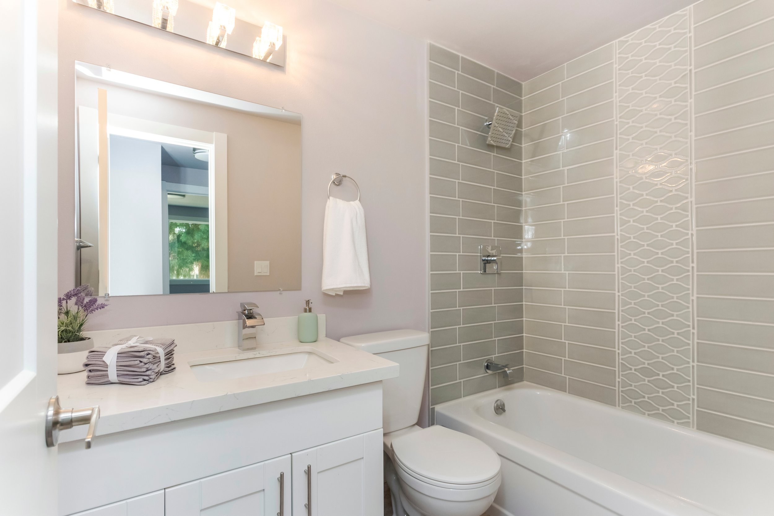 Modern bathroom with a white vanity, large mirror, and a bathtub with decorative tiling on the wall.