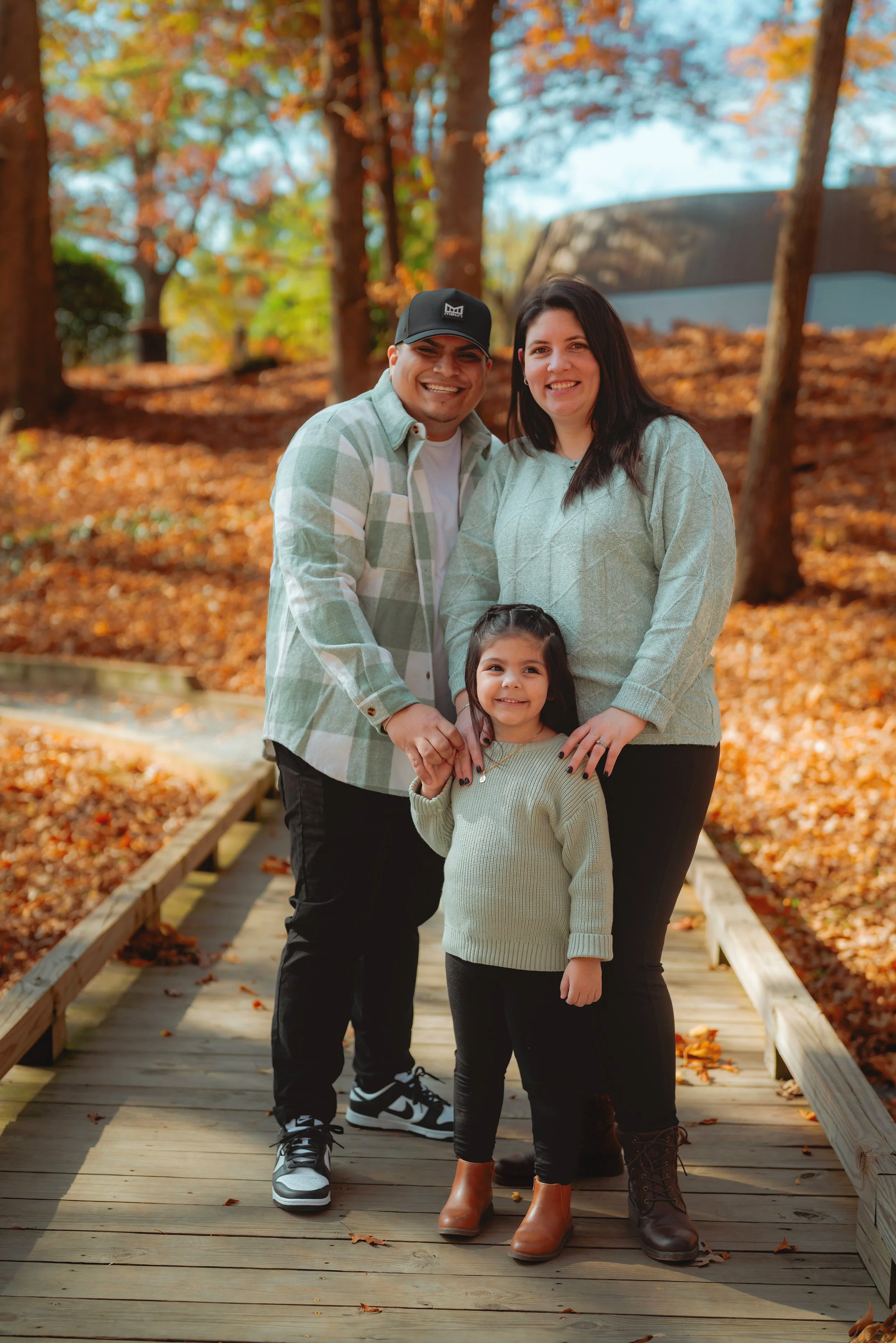 A family of three standing on a wooden bridge in a park during autumn, surrounded by fall foliage.