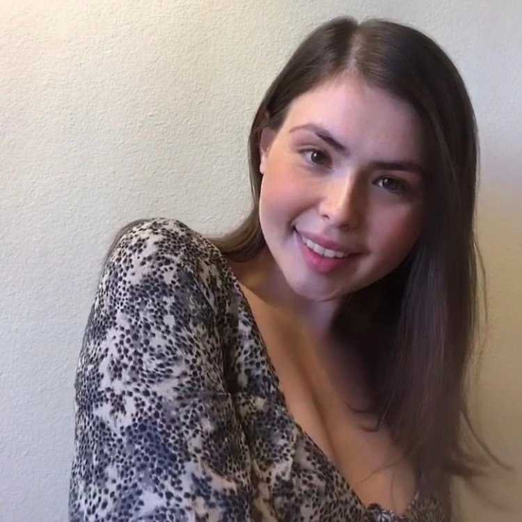 A young woman with long, straight brown hair smiling at the camera, wearing a leopard print top, standing against a plain light-colored wall.