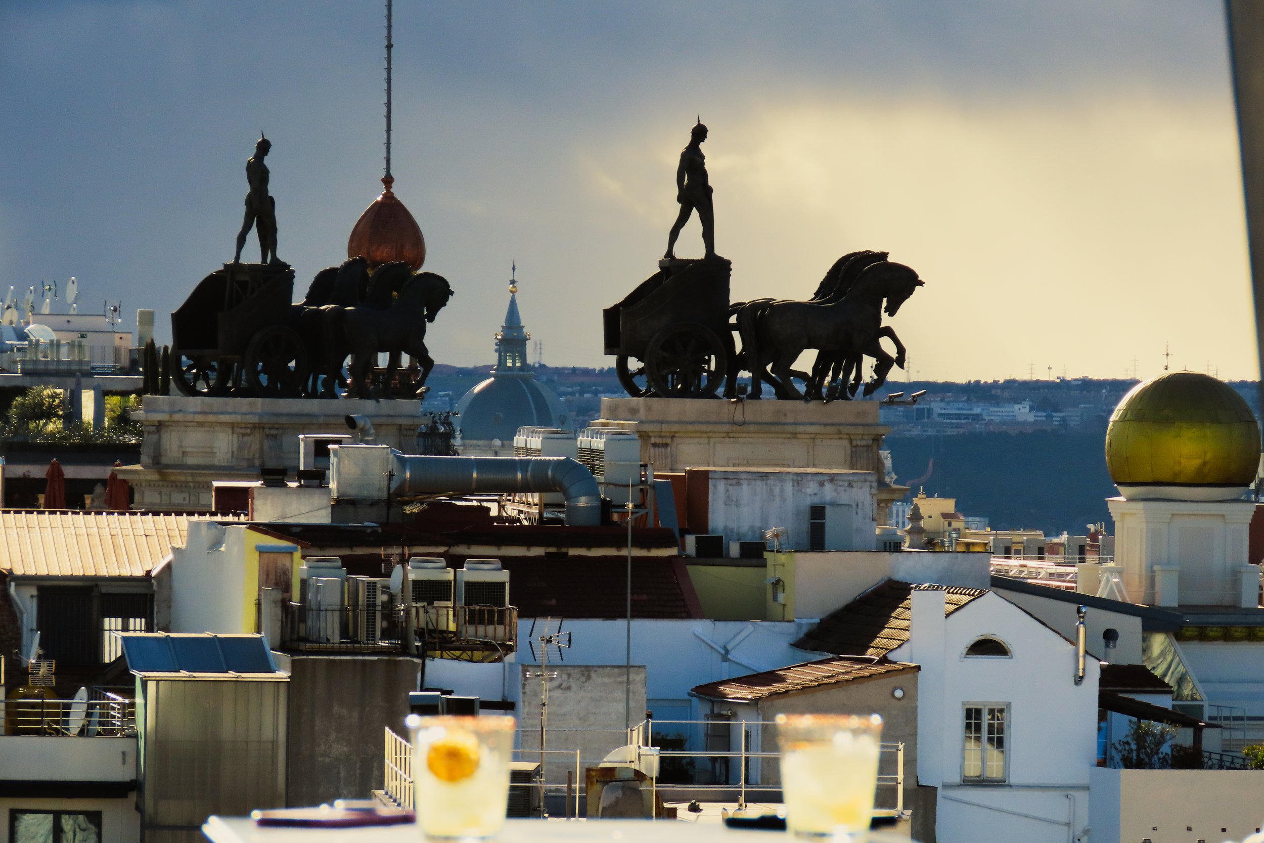 Vista di un tetto di una città, con statue di carri con cavalli e figure umane sul tetto. Sullo sfondo si vede la città e il cielo al tramonto.