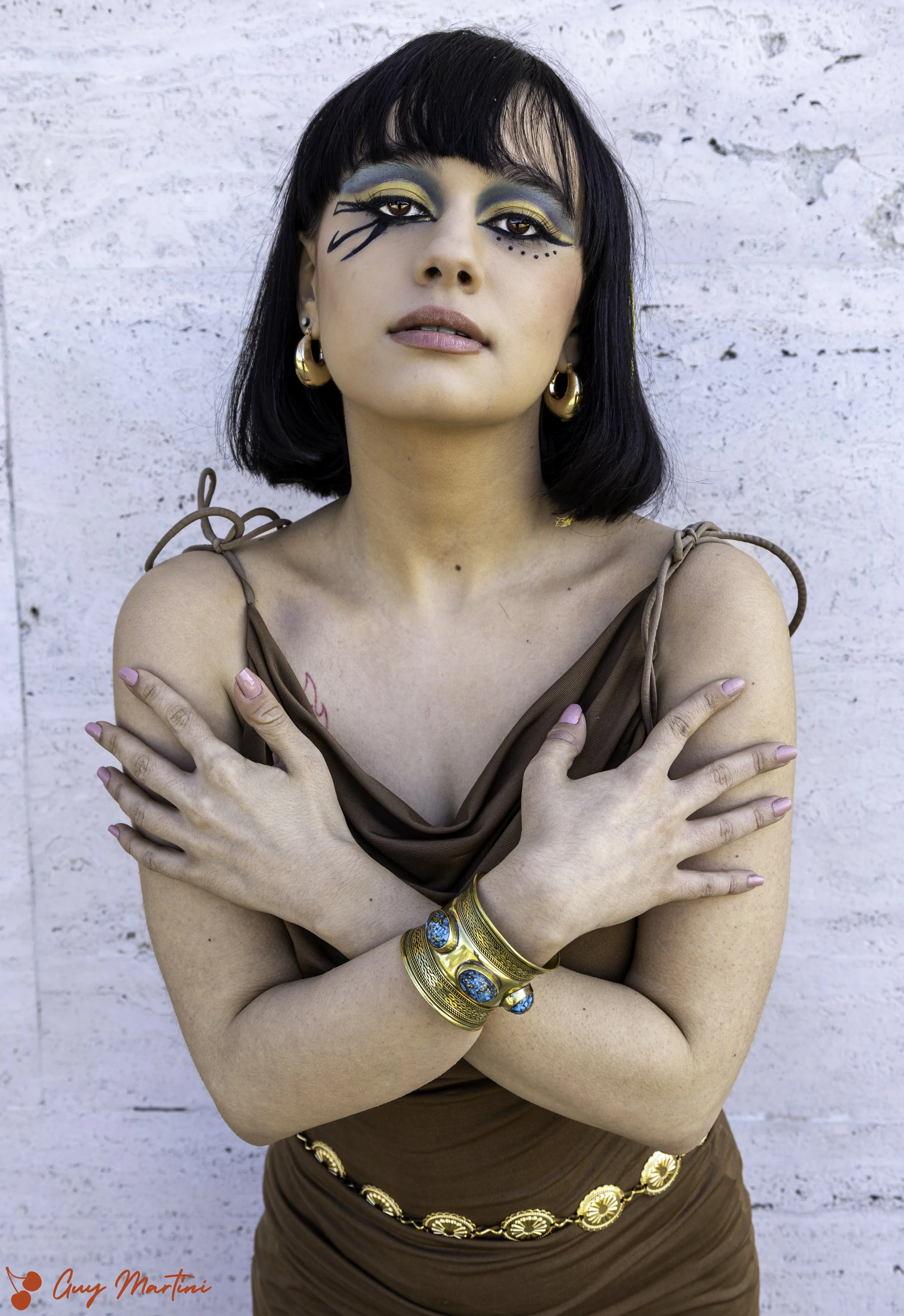 Donna con trucco elaborato e bracciale d'oro, posa con le braccia incrociate sul petto contro uno sfondo di parete bianca.