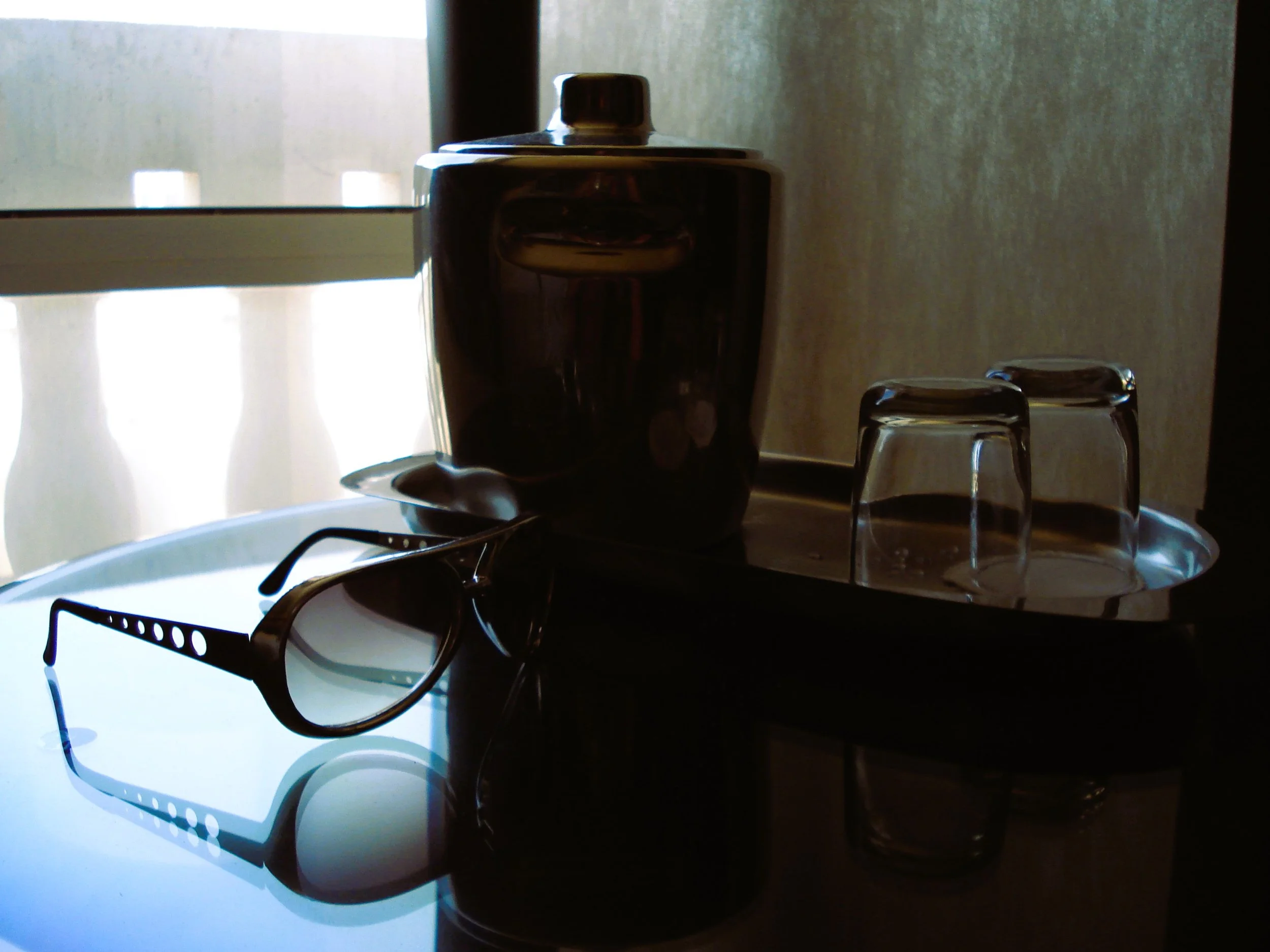 Occhiali da sole, lampada di vetro e bicchieri trasparenti su un vassoio, in un ambiente con luce naturale.