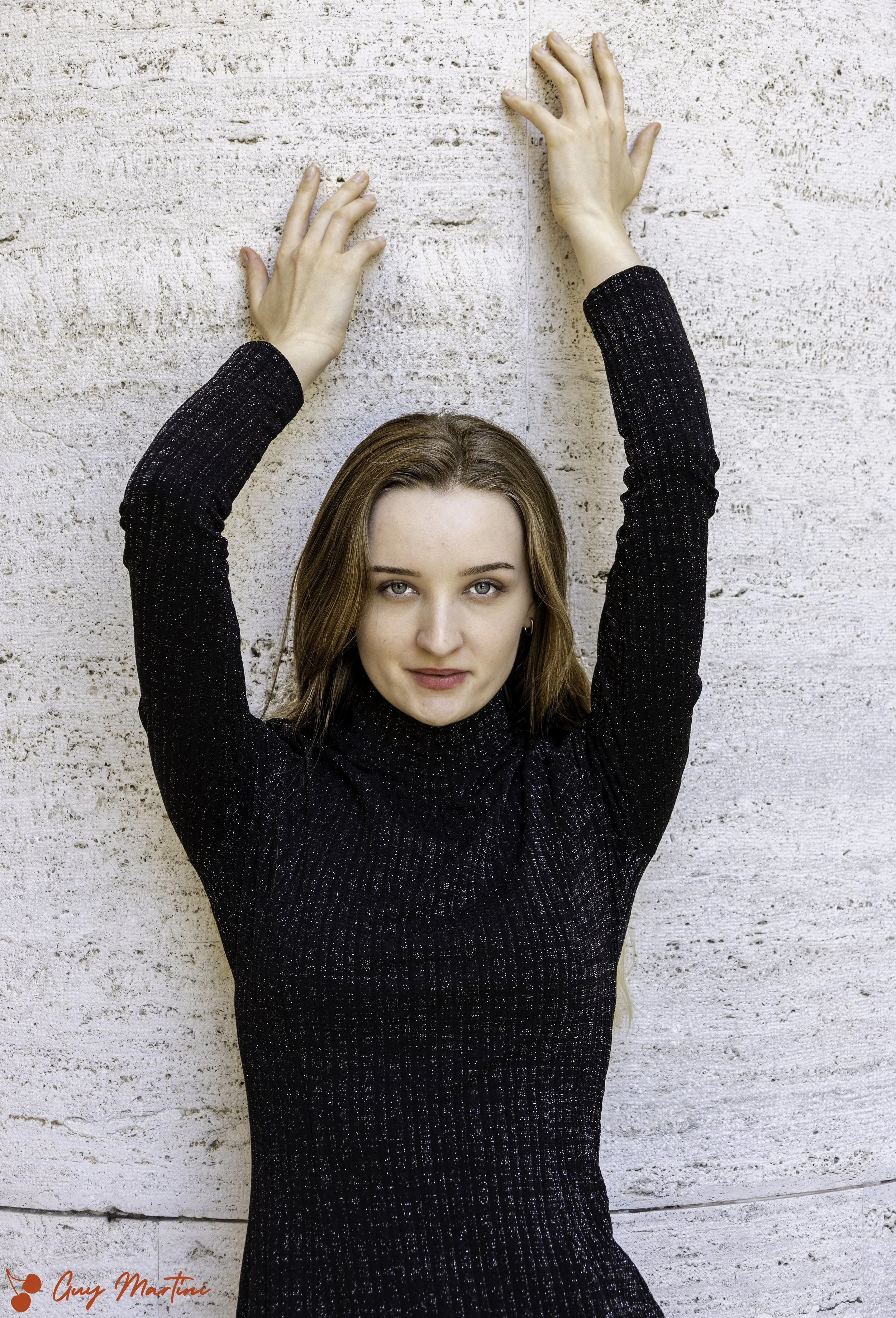Ragazza con capelli castani, che indossa un dolcevita nero con dettagli luminosi, si appoggia a una parete di pietra bianca, sollevando le braccia sopra la testa.