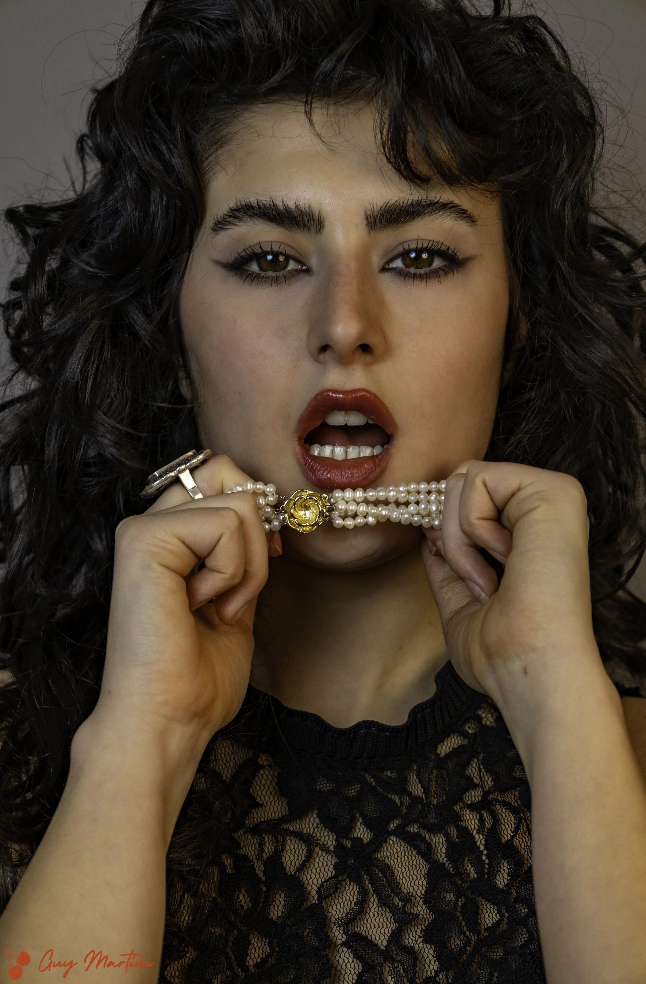 Una donna con capelli ricci, trucco marcato, che tiene una collana di perle con una chiusura dorata, con la bocca leggermente aperta, che sembra sorpresa o seducente.