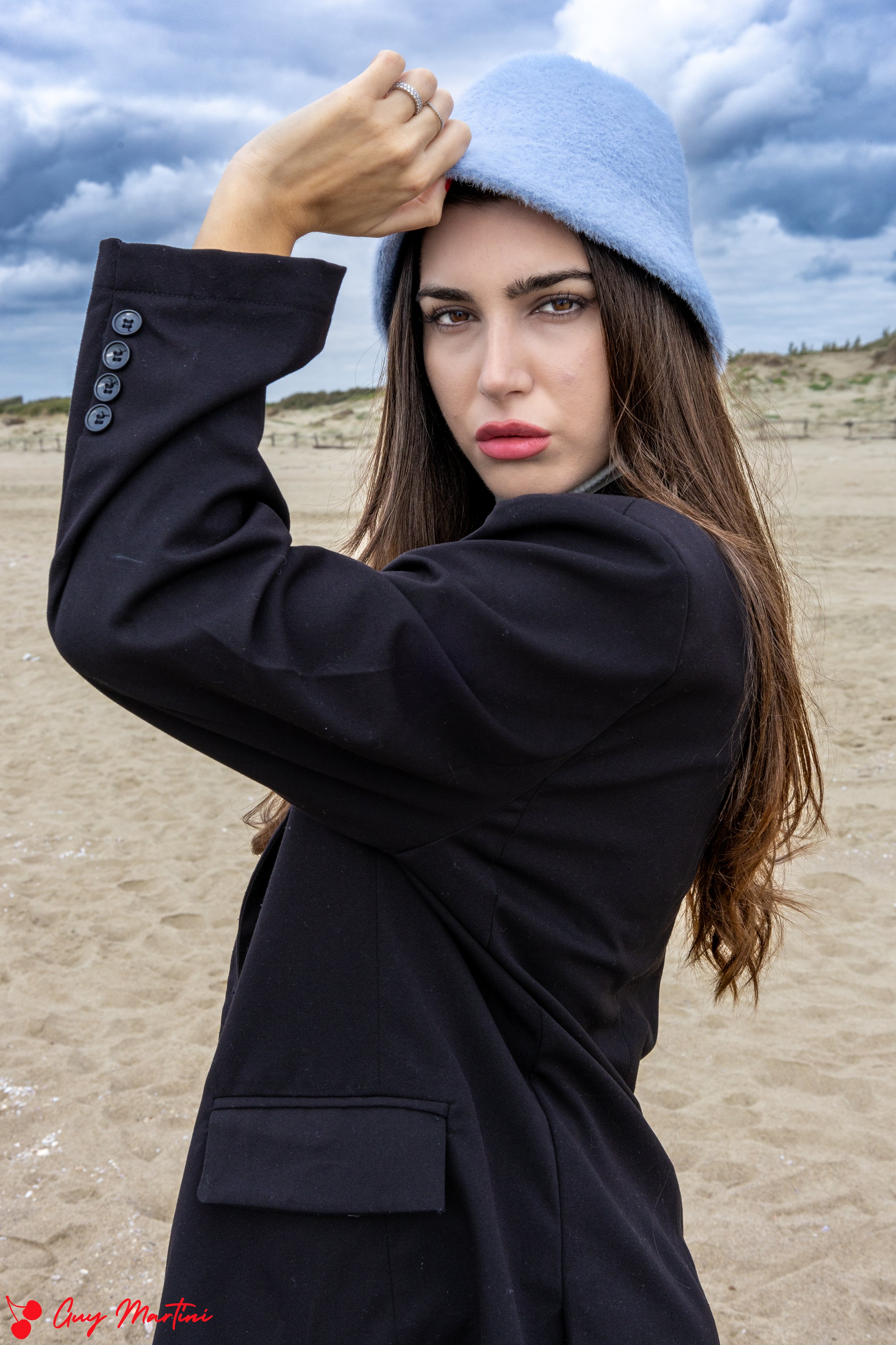 Ragazza con cappello azzurro indossa un cappotto nero in spiaggia con cielo nuvoloso.