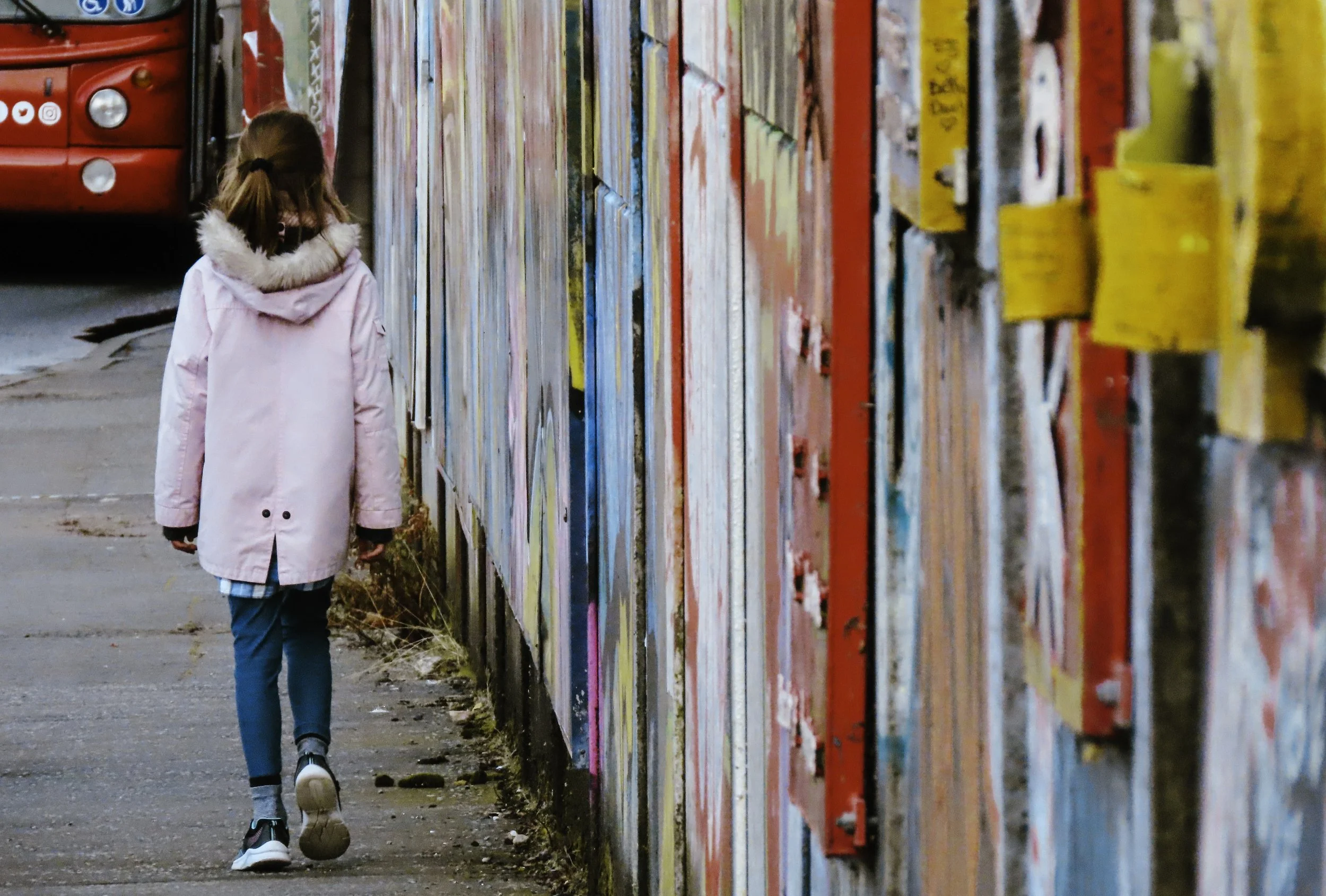 Una bambina con cappotto rosa e pantaloni blu cammina lungo un muro decorato con graffiti colorati.