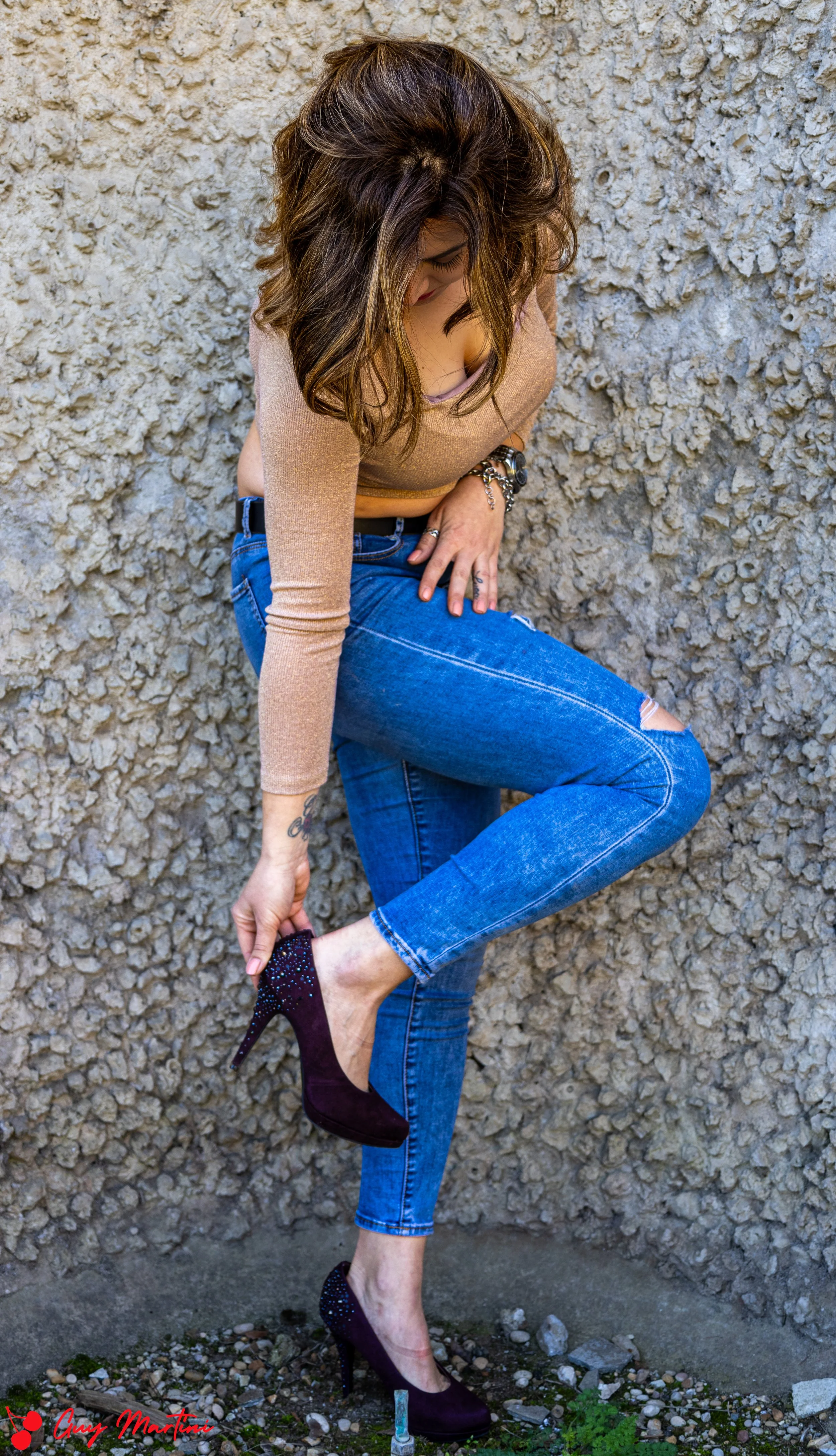 Donna con jeans blu, maglietta a maniche lunghe beige e tacchi neri con decorazioni brillante, si sta aggiustando la calzatura in un angolo di muro in cemento.