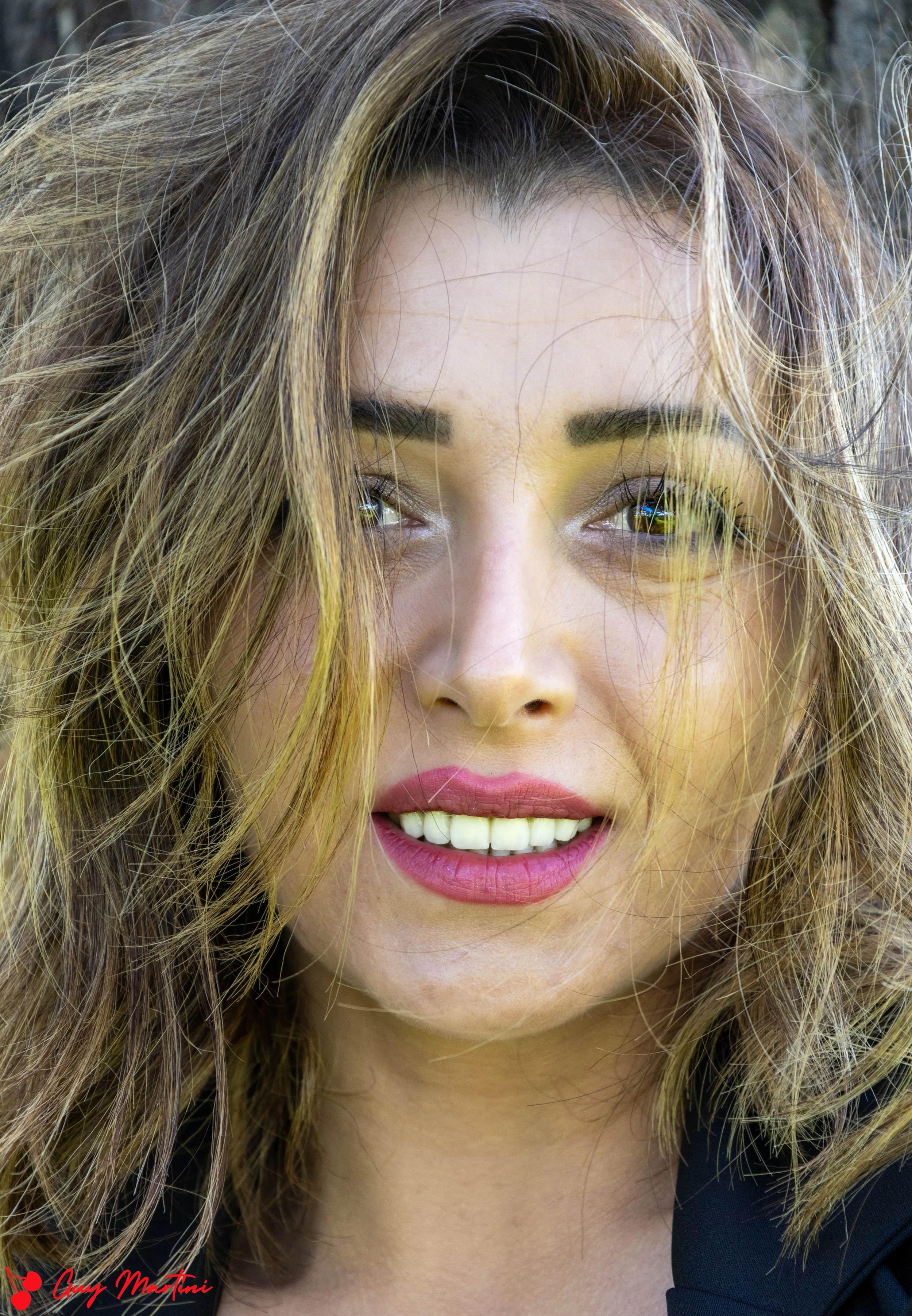 Primo piano di una donna con capelli ondulati, sorriso, labbra color rosa, e occhi chiari, sfondo naturale.