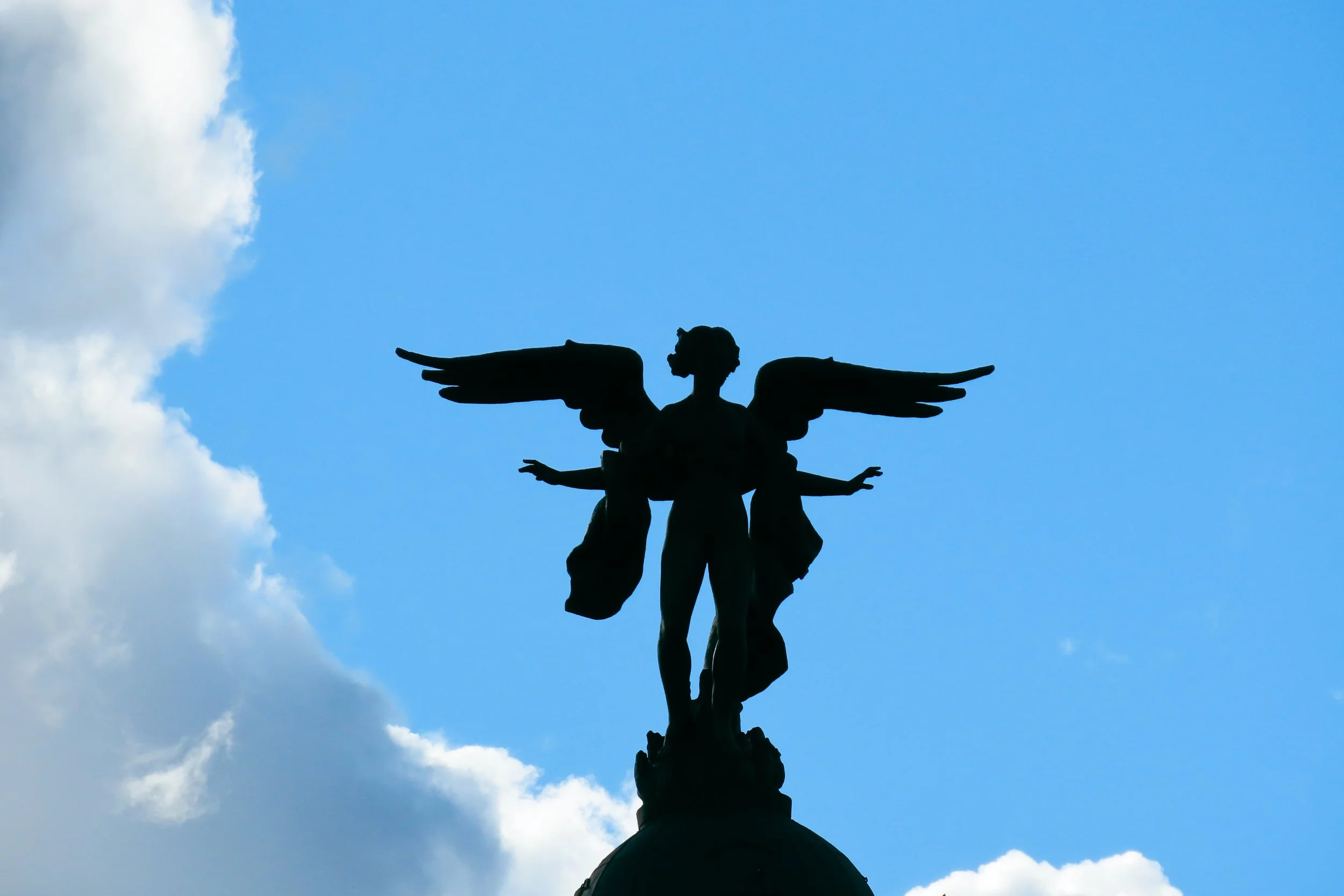 Statua di un angelo con ali estese contro il cielo azzurro con nuvole bianche.