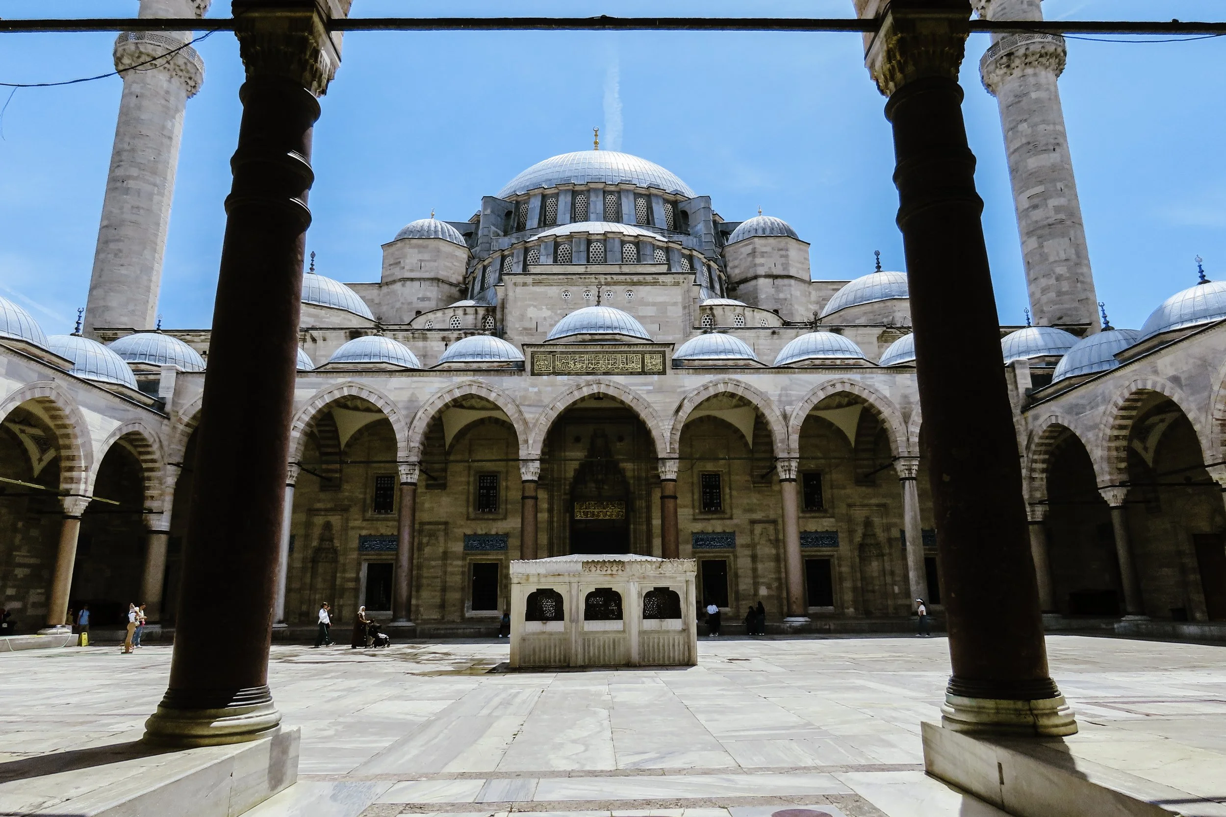 Vista dall'interno di una moschea con colonne di marmo e una grande cupola centrale, con un cortile aperto e alcuni visitatori.