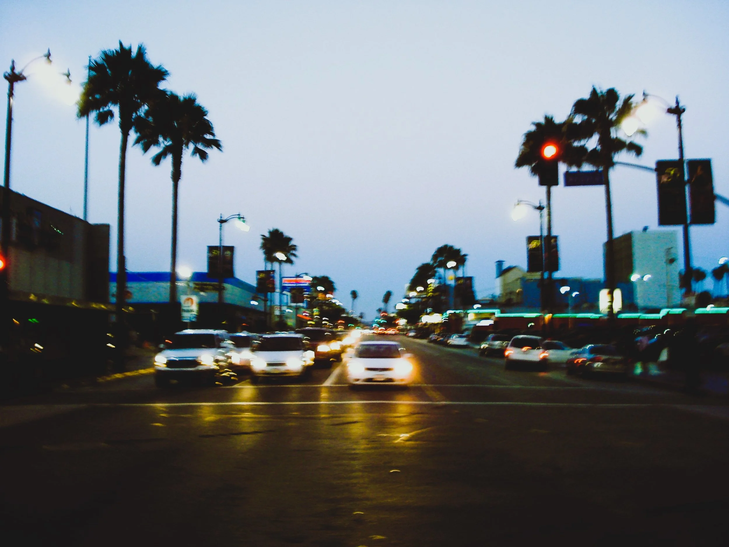 Strada trafficata con auto in movimento, palme lungo il marciapiede e luci di strada, al crepuscolo.
