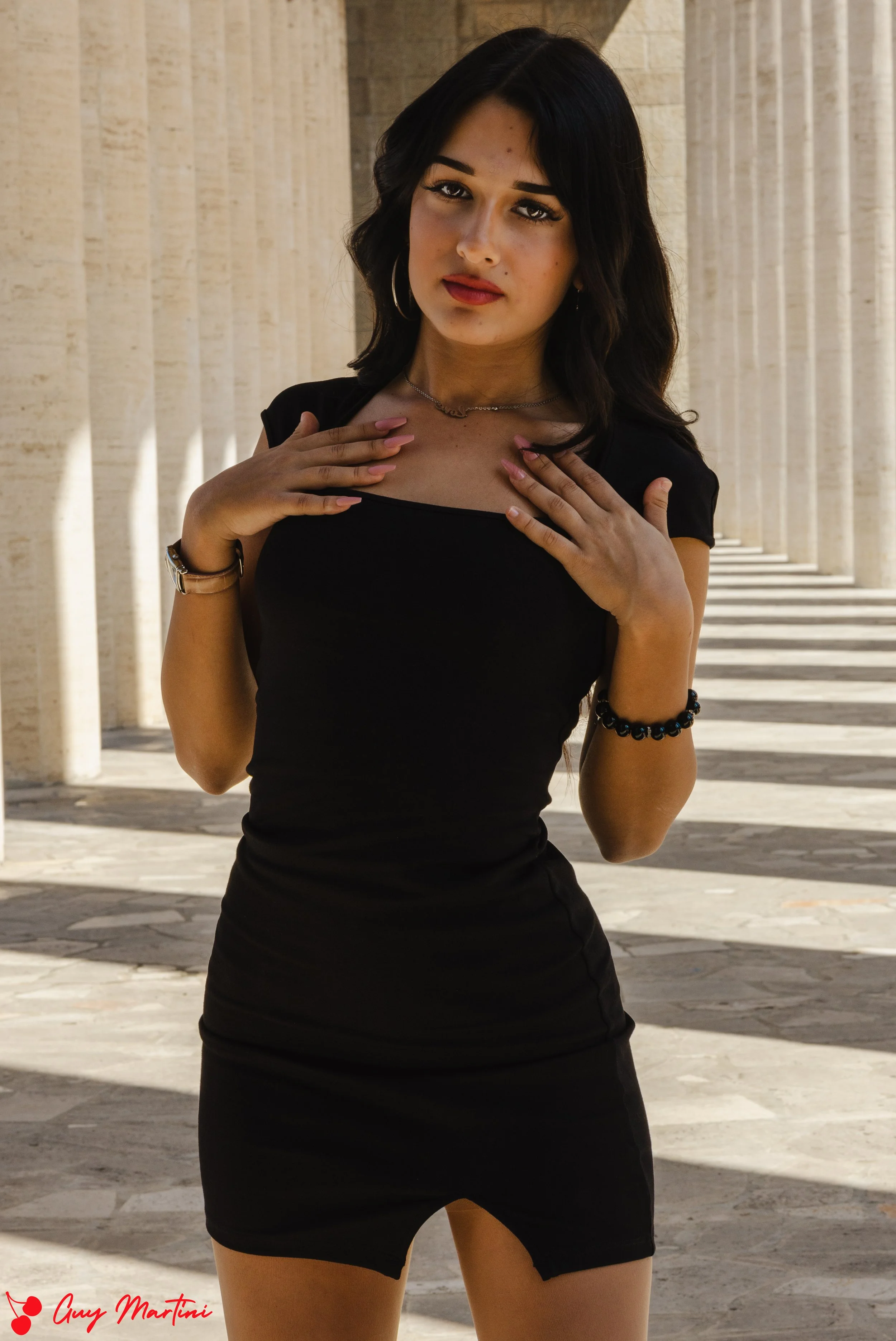 Una donna con capelli neri, indossa un vestito nero attillato e collane, si trova sotto una serie di colonne di pietra con luce che crea ombre sul pavimento.