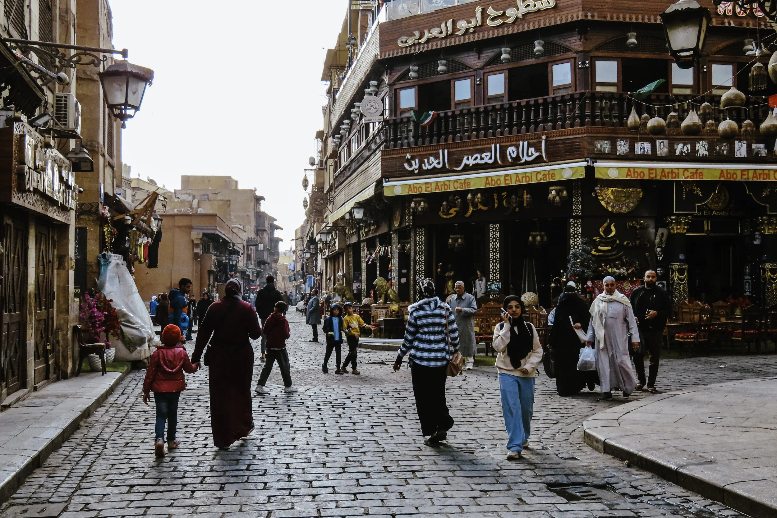Strada affollata con persone che camminano, negozi e ristorante con insegne in arabo, edifici di stile tradizionale.