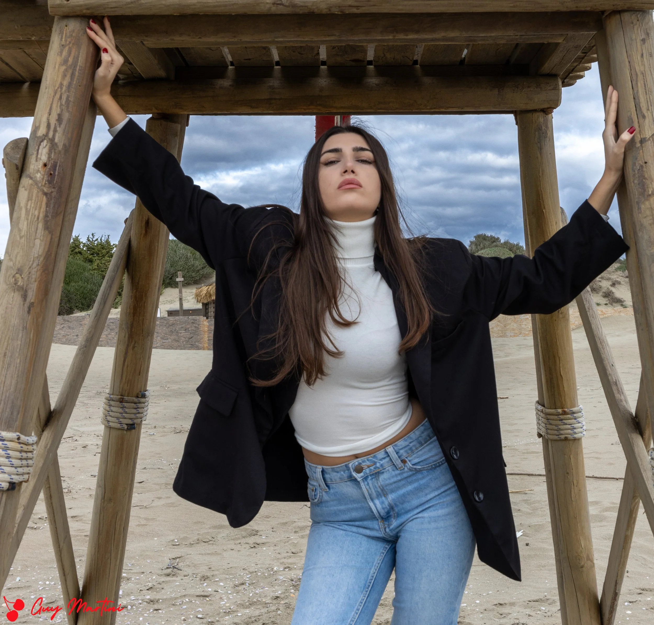 Una donna sta in piedi sotto una struttura di legno sulla spiaggia, con le braccia allargate che si appoggiano alle parti superiori dell'insieme, con un cielo nuvoloso sullo sfondo.