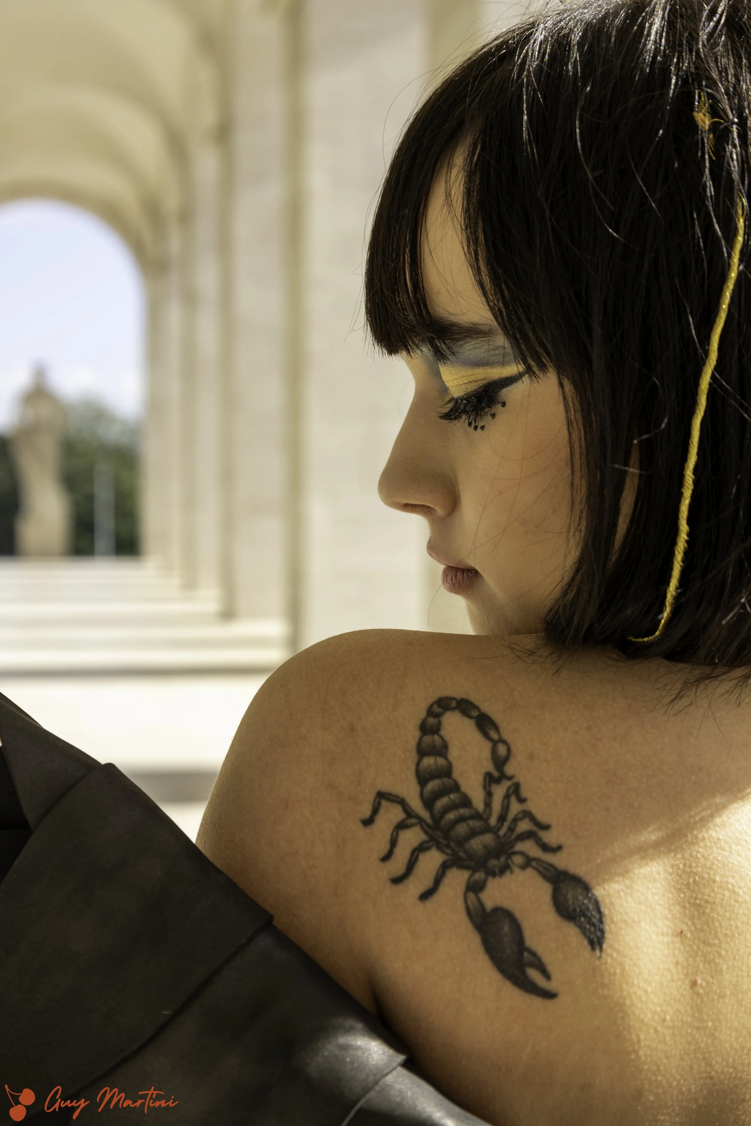 Donna con trucco artistico e capelli corti, che mostra un tatuaggio di uno scorpione sulla spalla