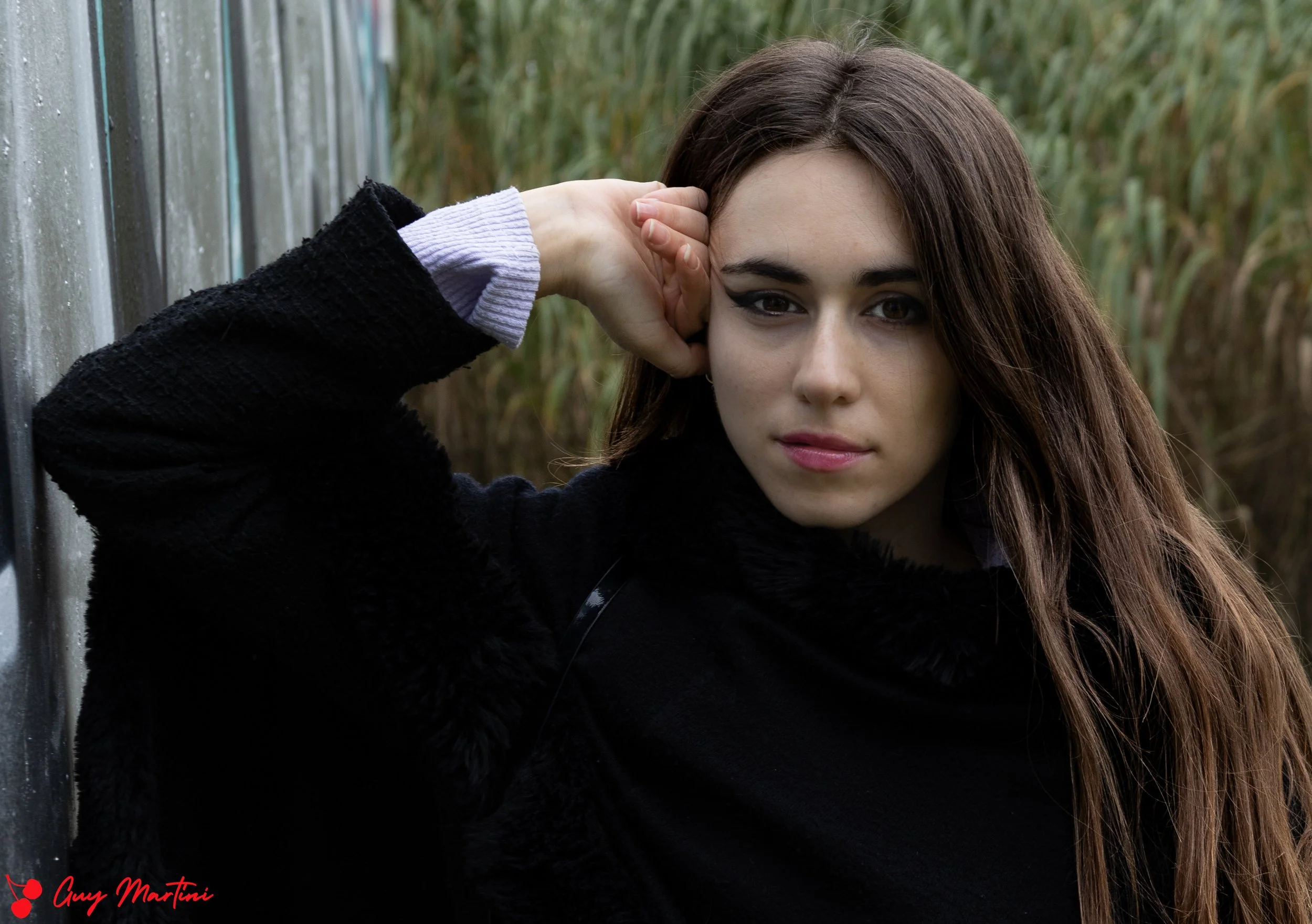 Una donna con capelli castani lunghi e trucco marcato posa con un'espressione seria accanto a una recinzione, in un ambiente naturale.