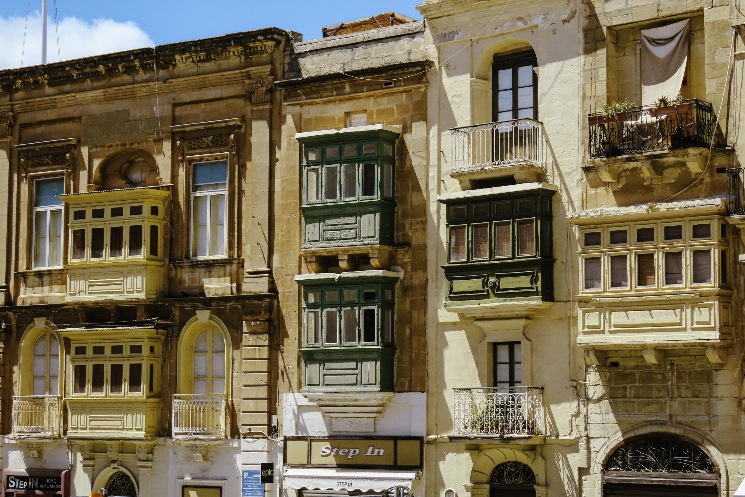 Edificio con balconi in stile tradizionale, alcune vetrate verdi e dettagli decorativi, sotto un cielo azzurro.