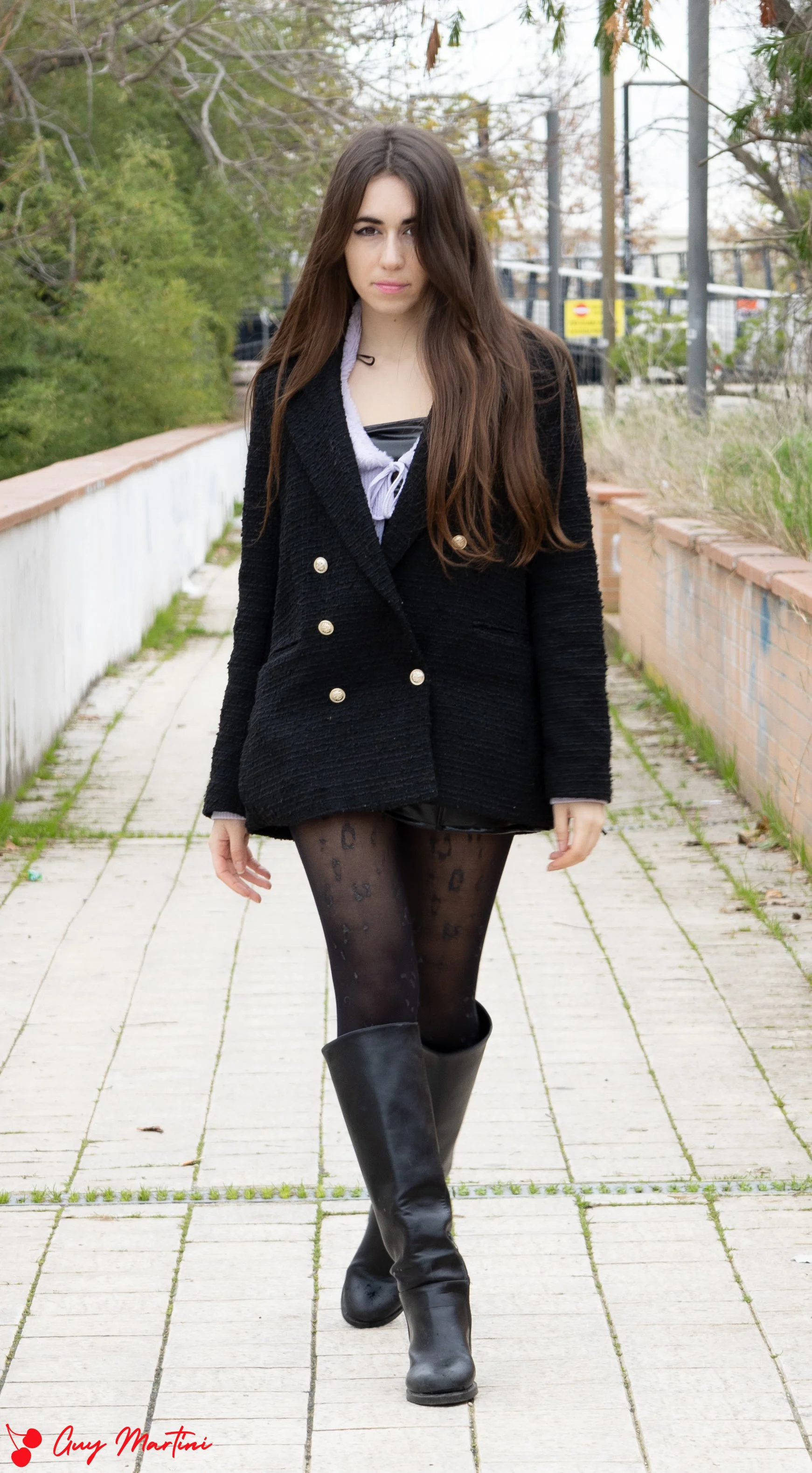 Una giovane donna cammina lungo un sentiero di cemento, indossando un cappotto nero, collant trasparenti con stampa leopardata, e stivali neri. gli alberi sullo sfondo hanno foglie verdi e marroni, indicando un cambio di stagione o autunno.