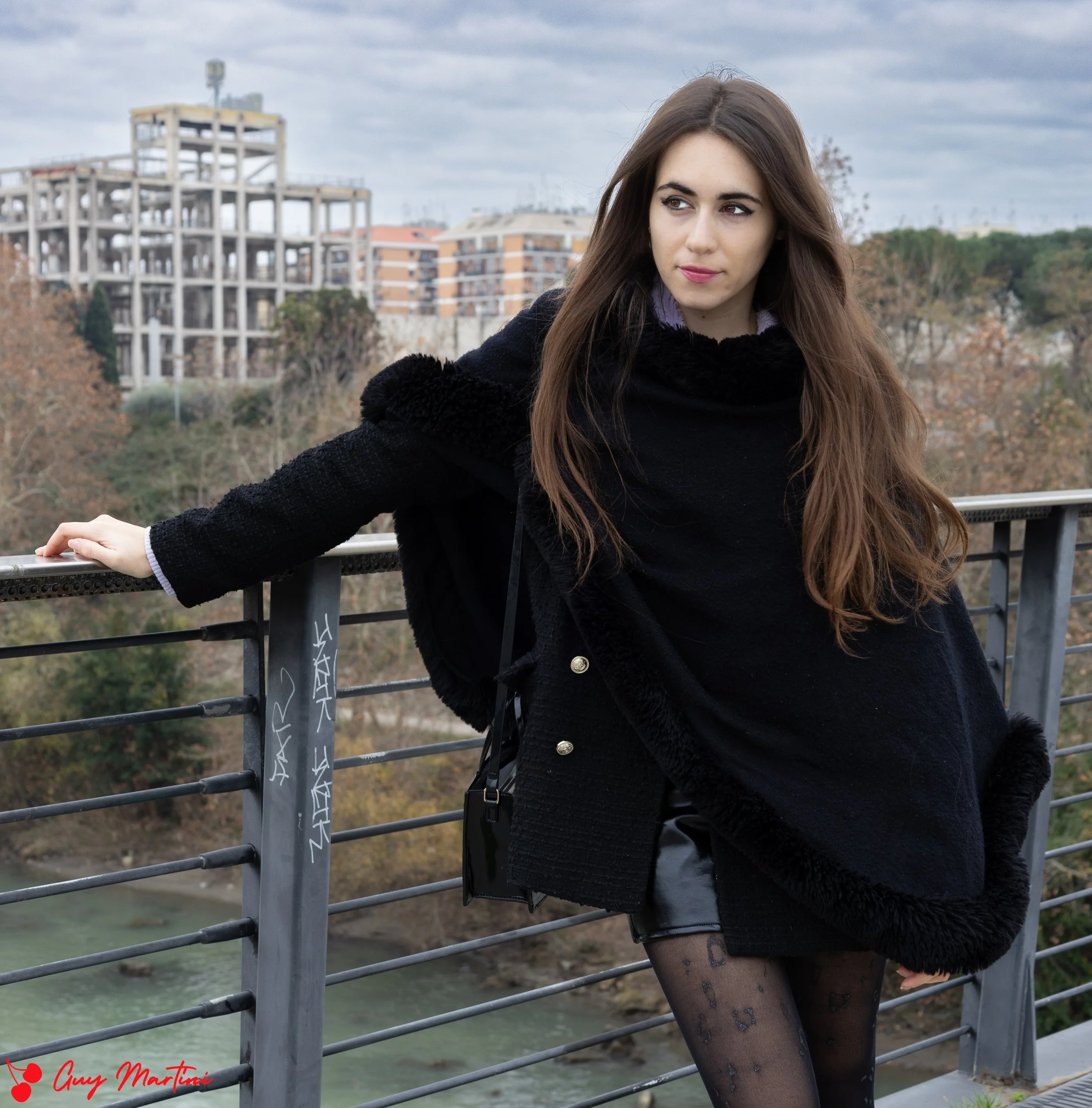 Una giovane donna con i capelli castani, vestita con un cappotto nero, si trova sul ponte con vista sul fiume e sulla città sullo sfondo con edifici in costruzione e alberi spogli. Sta appoggiando una mano sulla ringhiera.