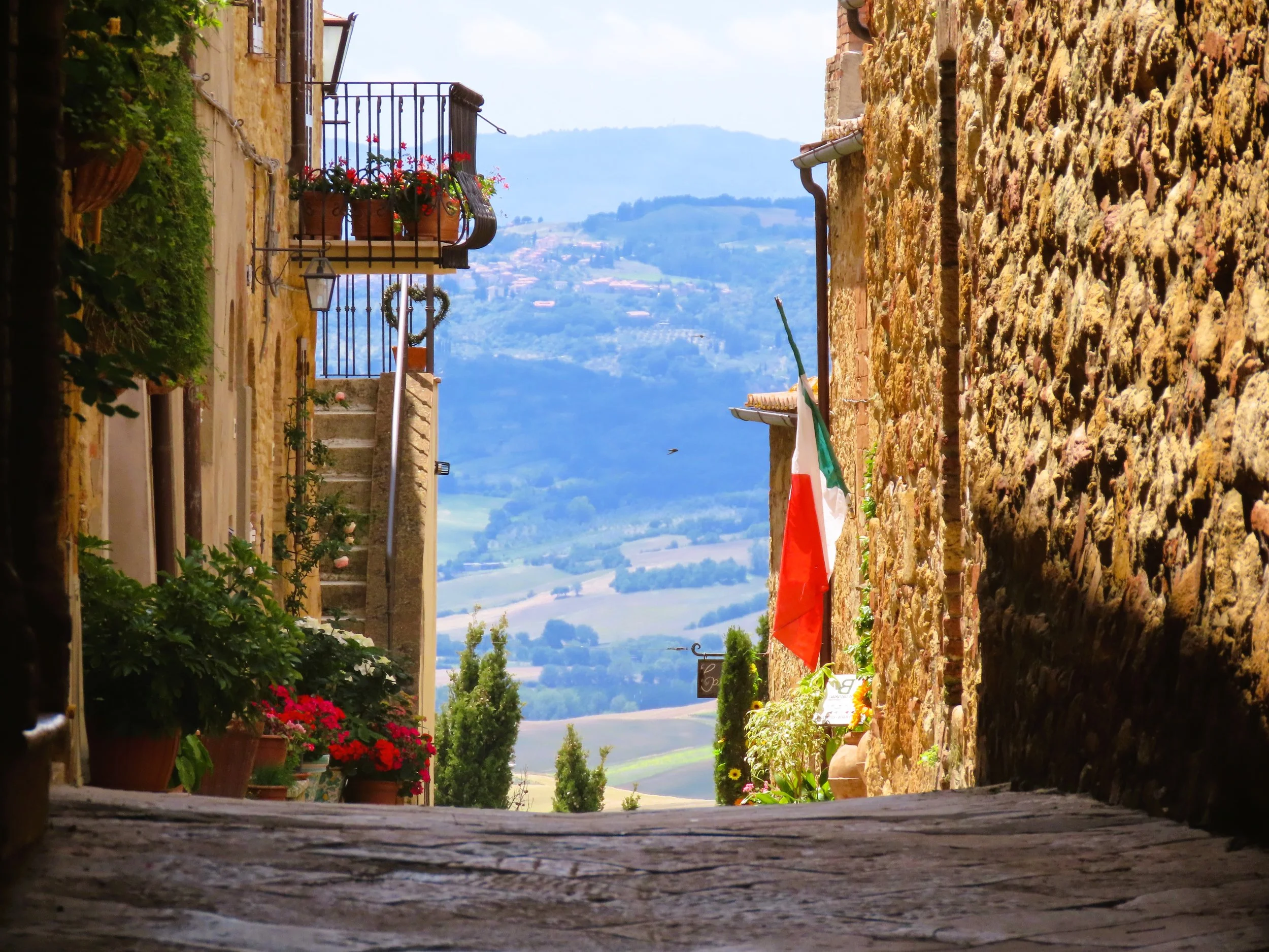 Vista di un vicolo in un paesino italiano con edifici in pietra, piante e fiori, bandiera italiana e vista sulle colline in lontananza.