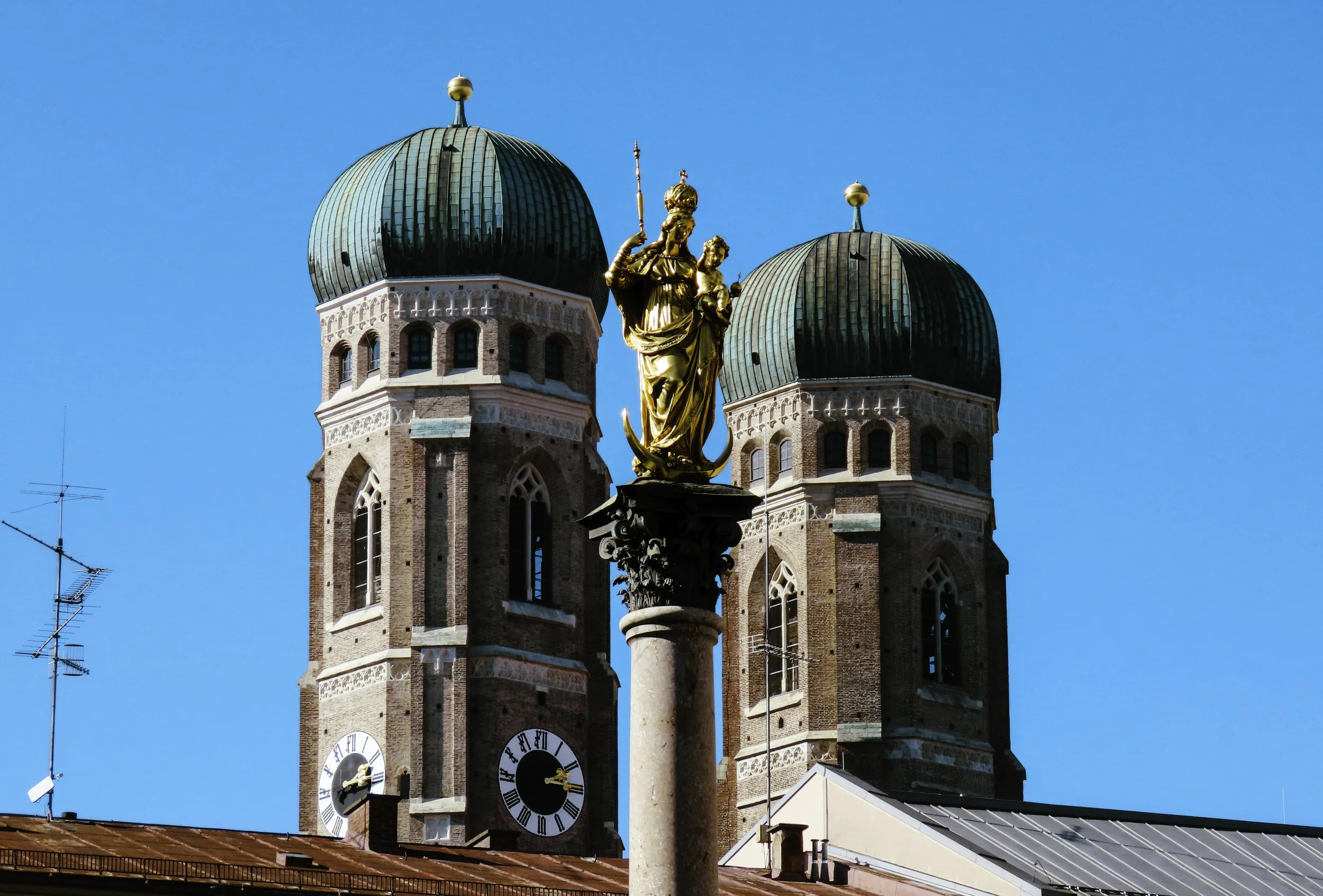 Statua dorata di una donna con bambino su una colonna, con due torri con cupole verdi e orologi sullo sfondo, sotto un cielo azzurro.