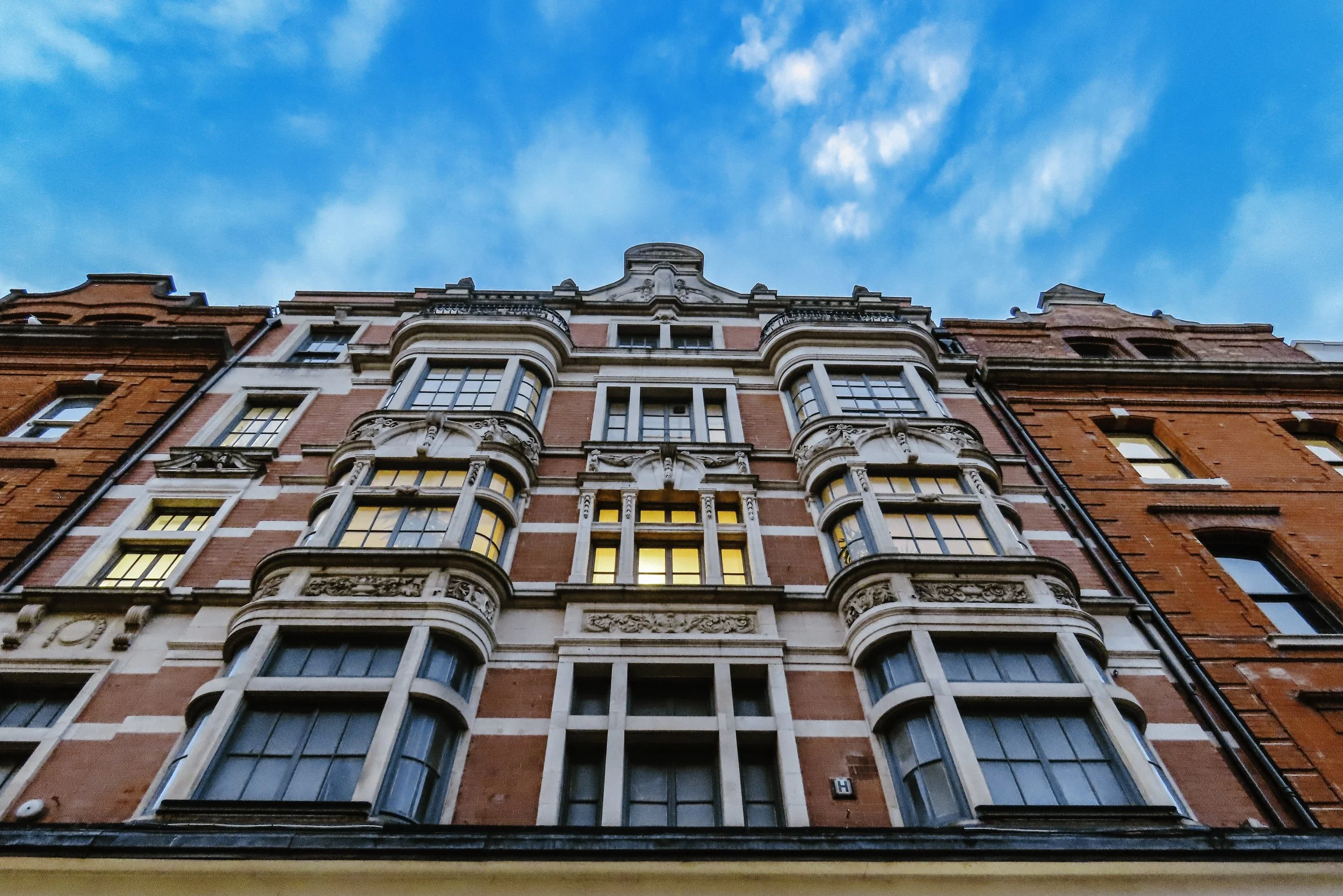 Edificio storico con facciata rossa e dettagli in pietra, finestre grandi e archi decorativi, cielo blu con nuvole sparse.