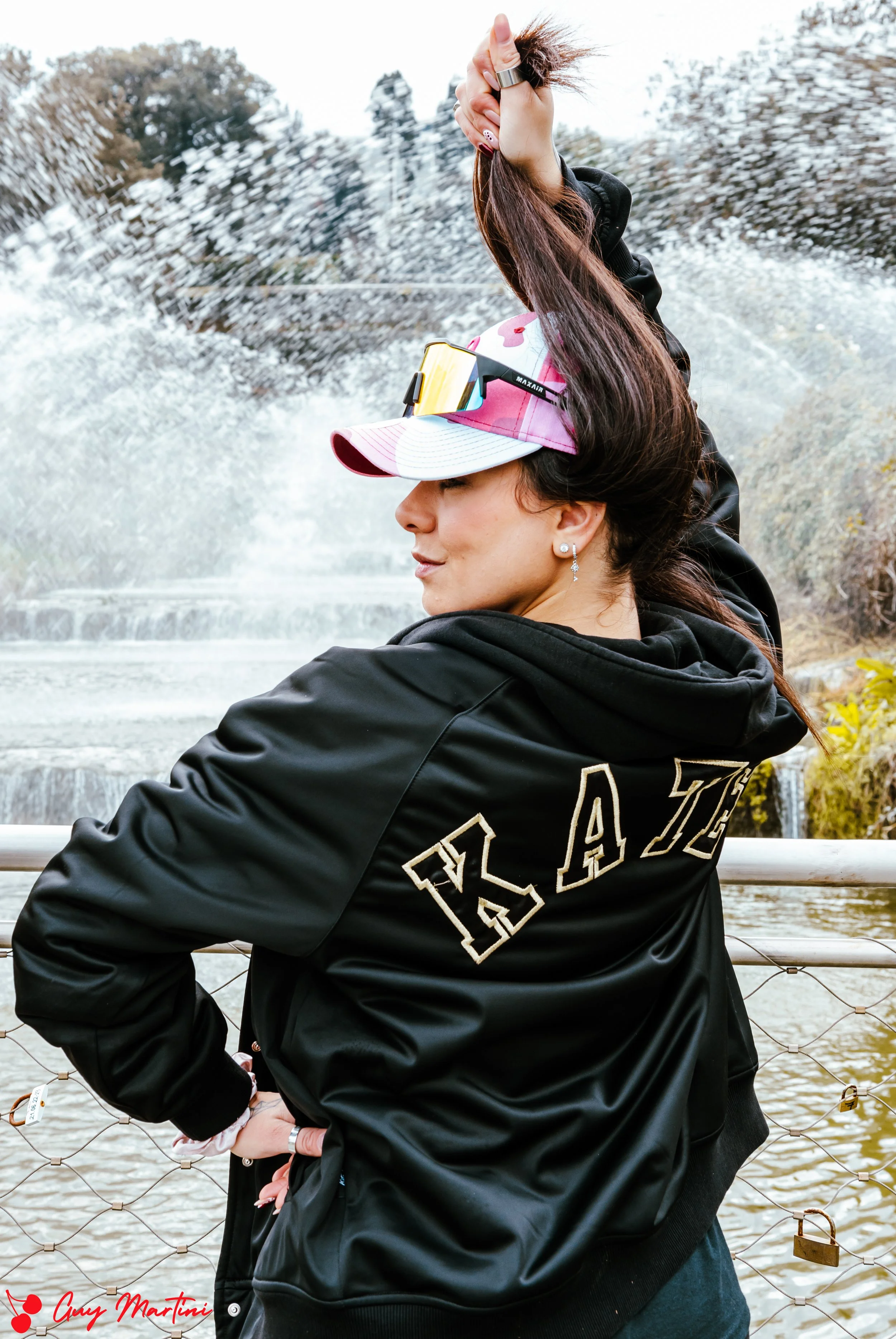 Una donna casual indossa una giacca patchwork con la scritta 'PLAZ' sulla schiena, cappello rosa con visiera, scarpe da ginnastica e occhiali da sole, mentre si trova vicino a una ringhiera con una cascata sullo sfondo.