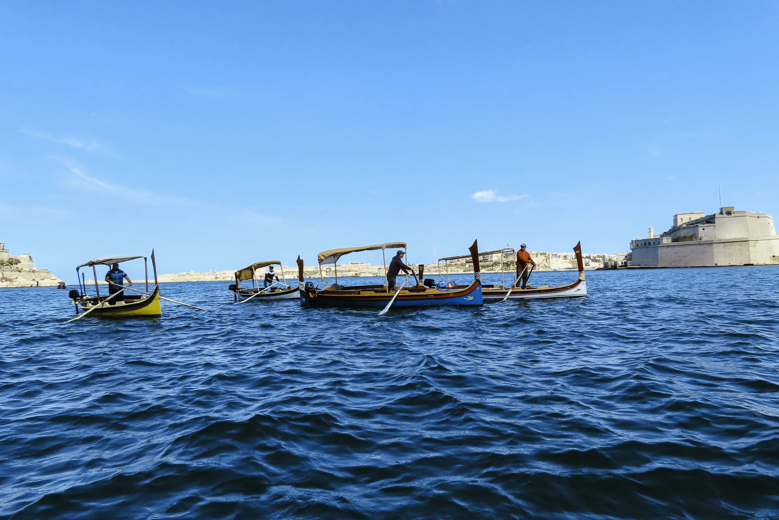 Quattro barche a remo in acque blu con uno sfondo di edifici storici di Malta sotto un cielo azzurro
