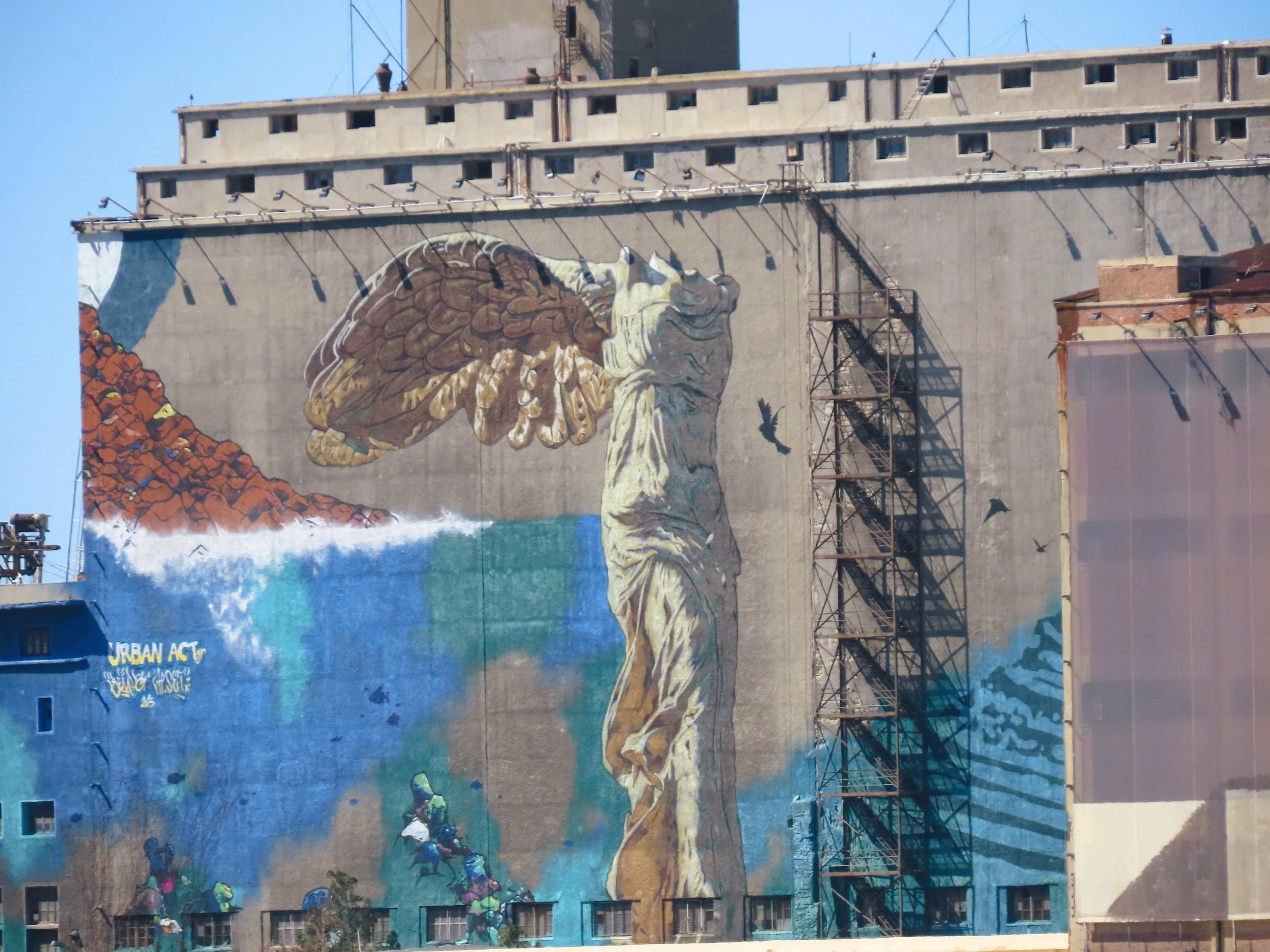 Murale che raffigura la statua della libertà con ali di uccelli e un paesaggio marino sullo sfondo, con alcuni graffiti e un'insegna 'URBAN ACT' in basso a sinistra.