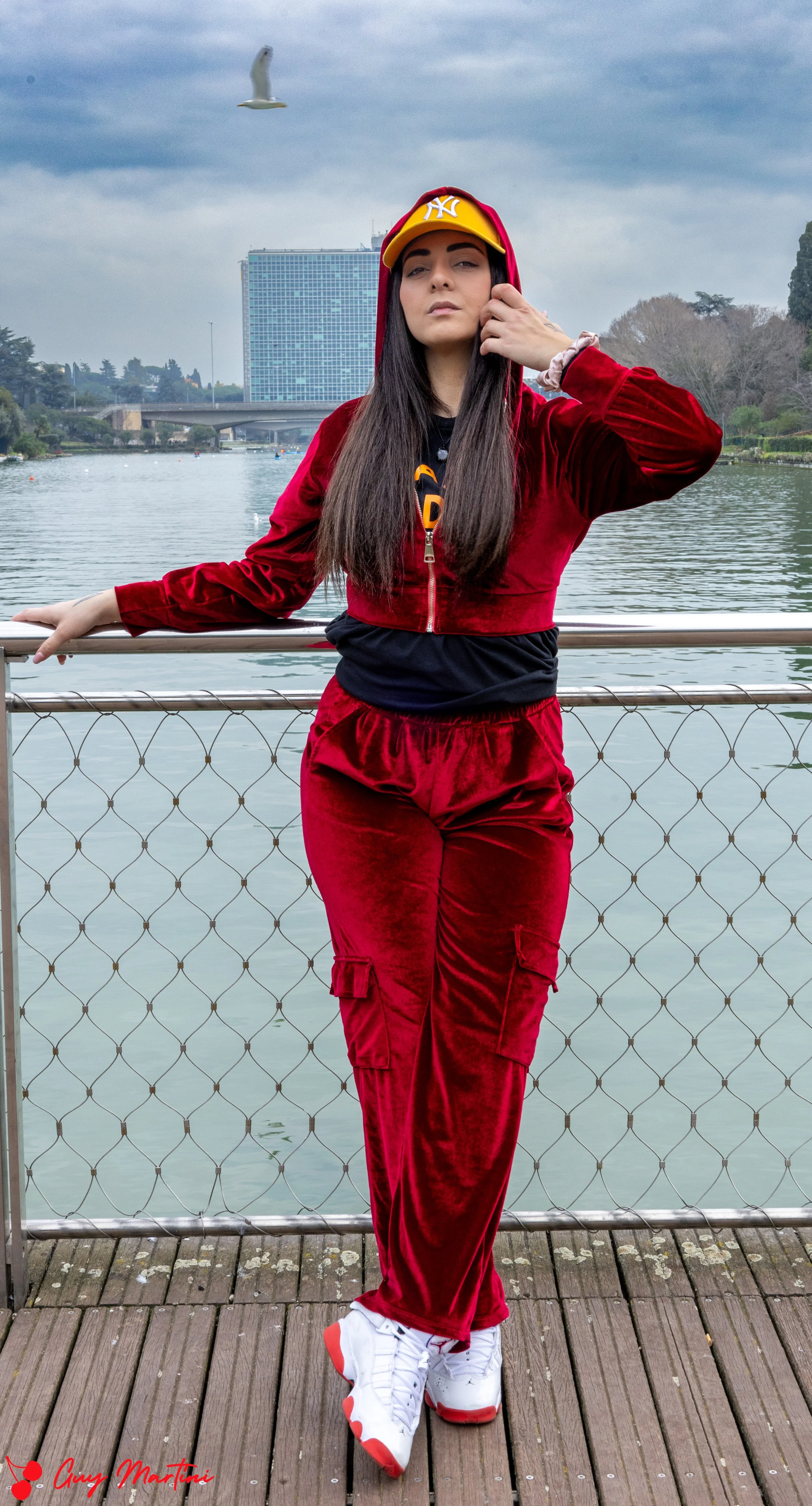 Una ragazza con un completo rosso in velluto, cappellino giallo con logo NY, scarpe bianche con dettagli rossi, si trova su un molo vicino a un lago o fiume, con edifici e un cielo nuvoloso sullo sfondo.