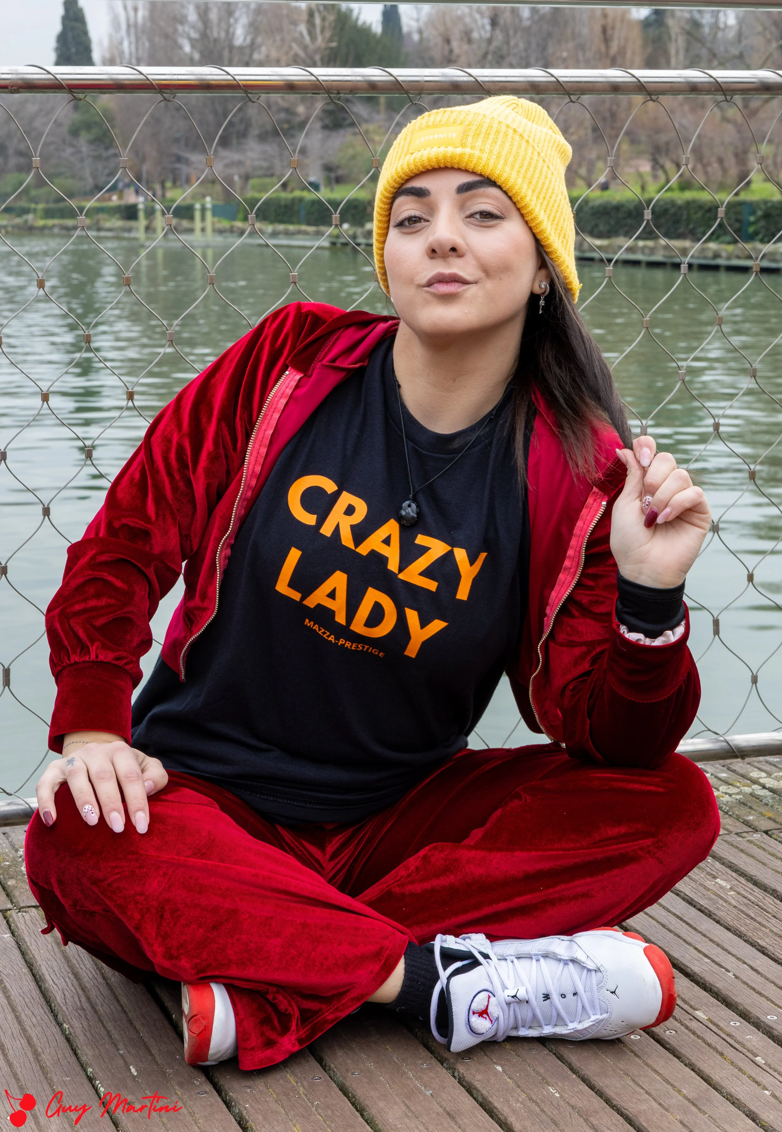Una donna seduta a gambe incrociate su una piattaforma di legno vicino a un lago, indossa un cappellino giallo, una felpa rossa e una maglietta nera con la scritta 'CRAZY LADY' di colore arancione. Sul lato inferiore dell'immagine c'è una firma rossa