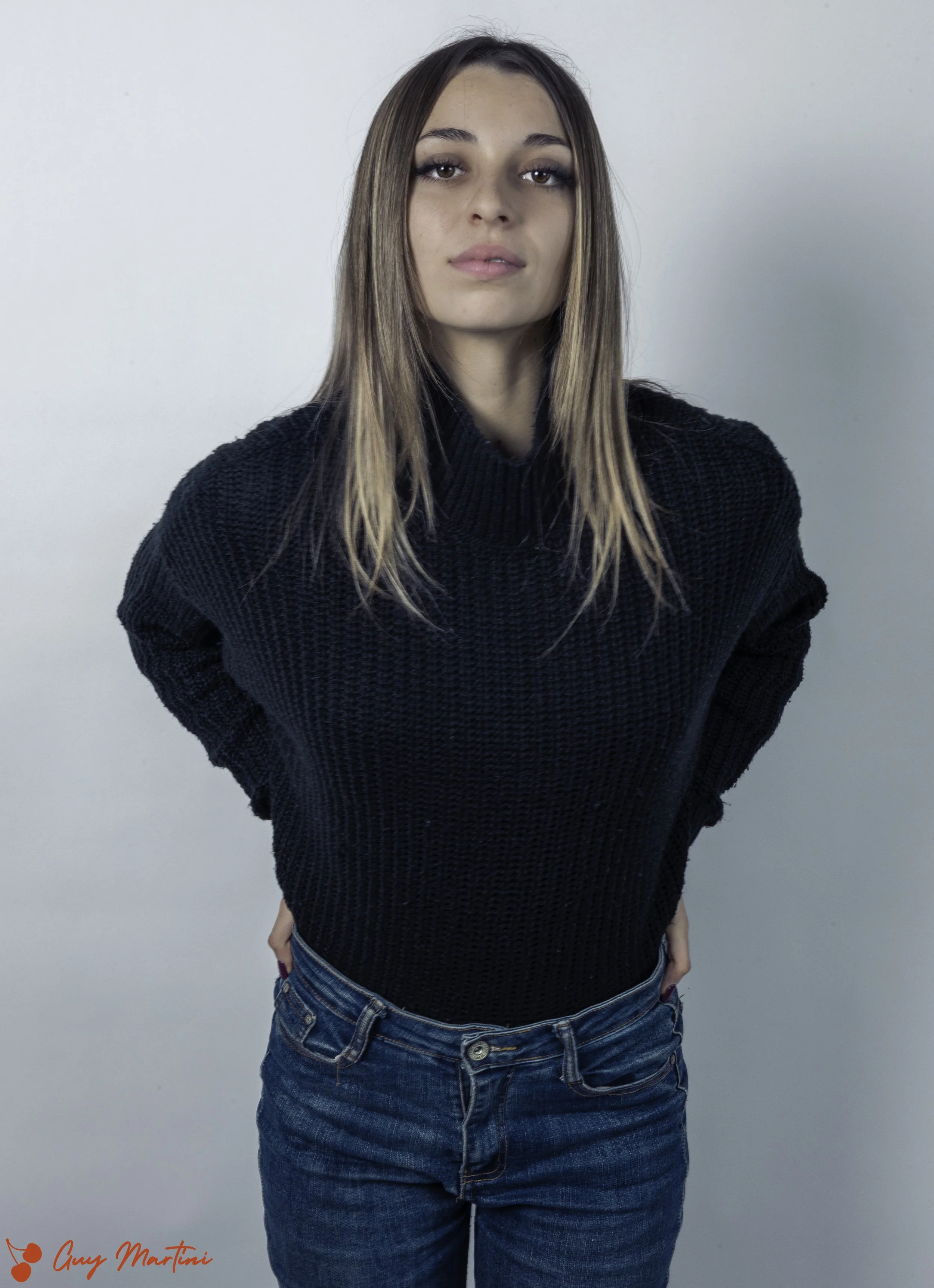 Donna giovane con capelli castani lunghi, indossa maglione nero e jeans, sfondo bianco.