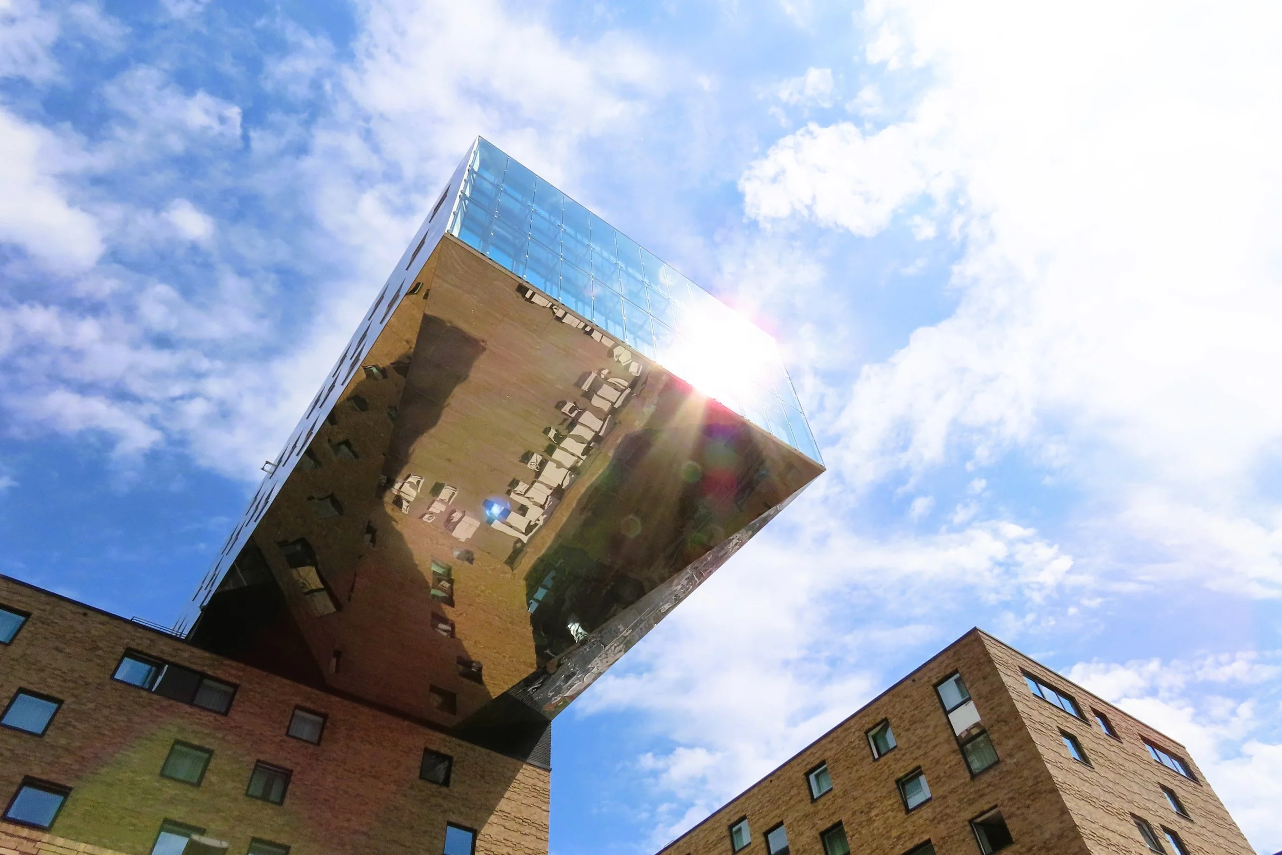Edificio moderno con facciata di vetro e metallo, riflettendo le nuvole e i bambini sottostanti, sotto un cielo azzurro con nuvole sparse.