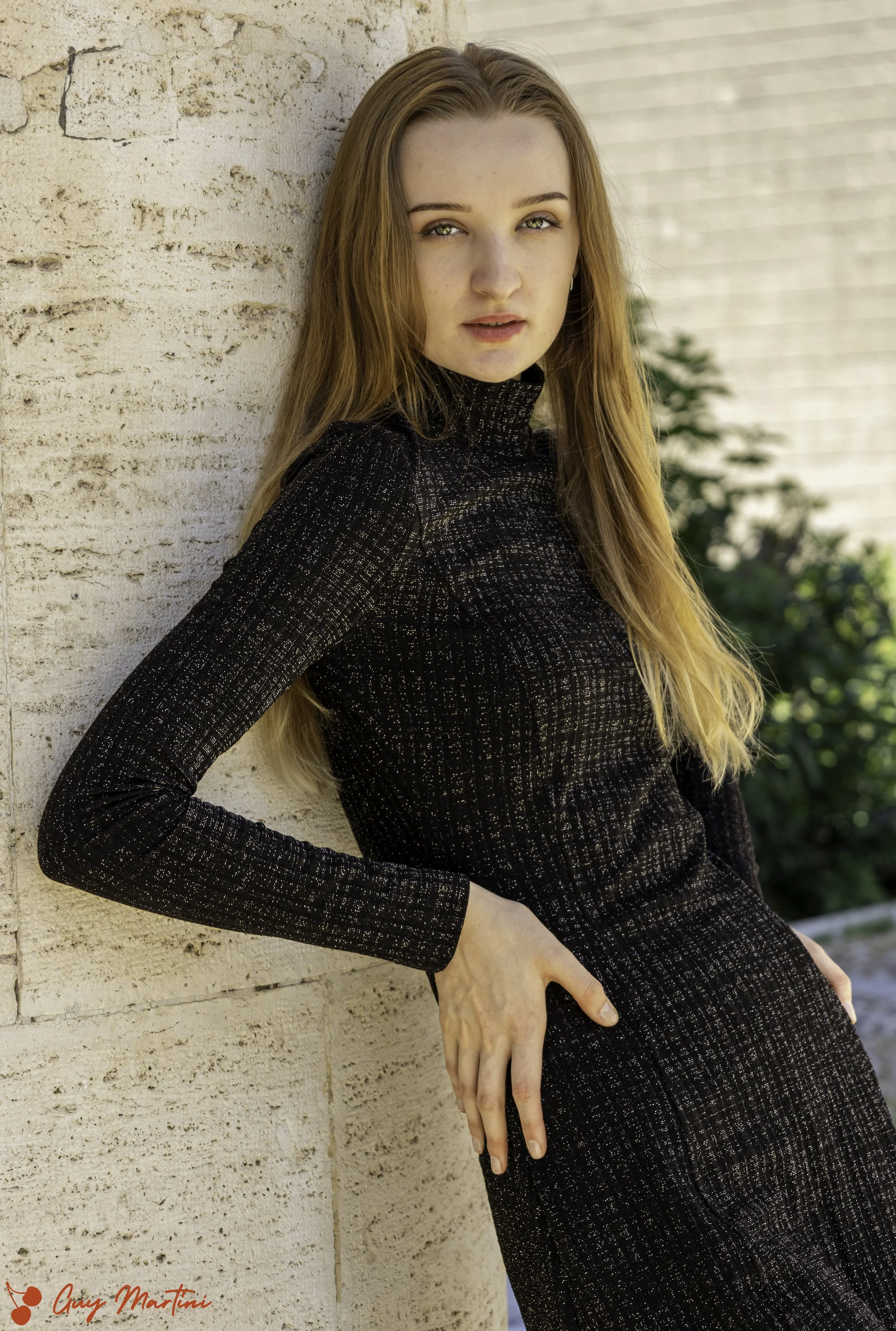 Ragazza con capelli lunghi e castani, indossa un vestito nero gli dettagli lucidi, si appoggia a una parete di mattoni chiara in un ambiente esterno ben illuminato.