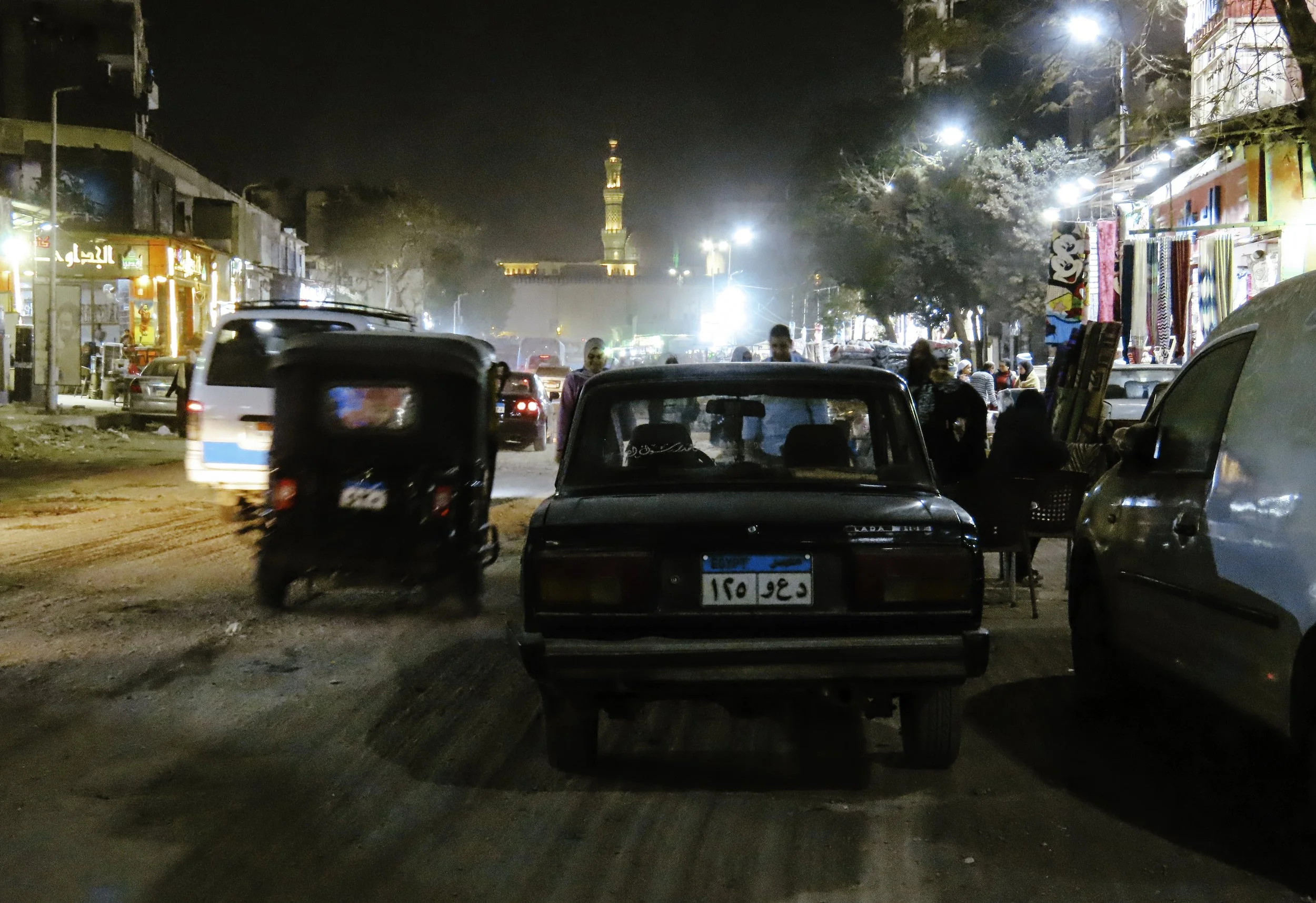 Una strada urbana di sera con automobili, motocicli e venditori ambulanti. Sullo sfondo si vede una torre illuminata e alcune bancarelle con stoffe e altre merci.