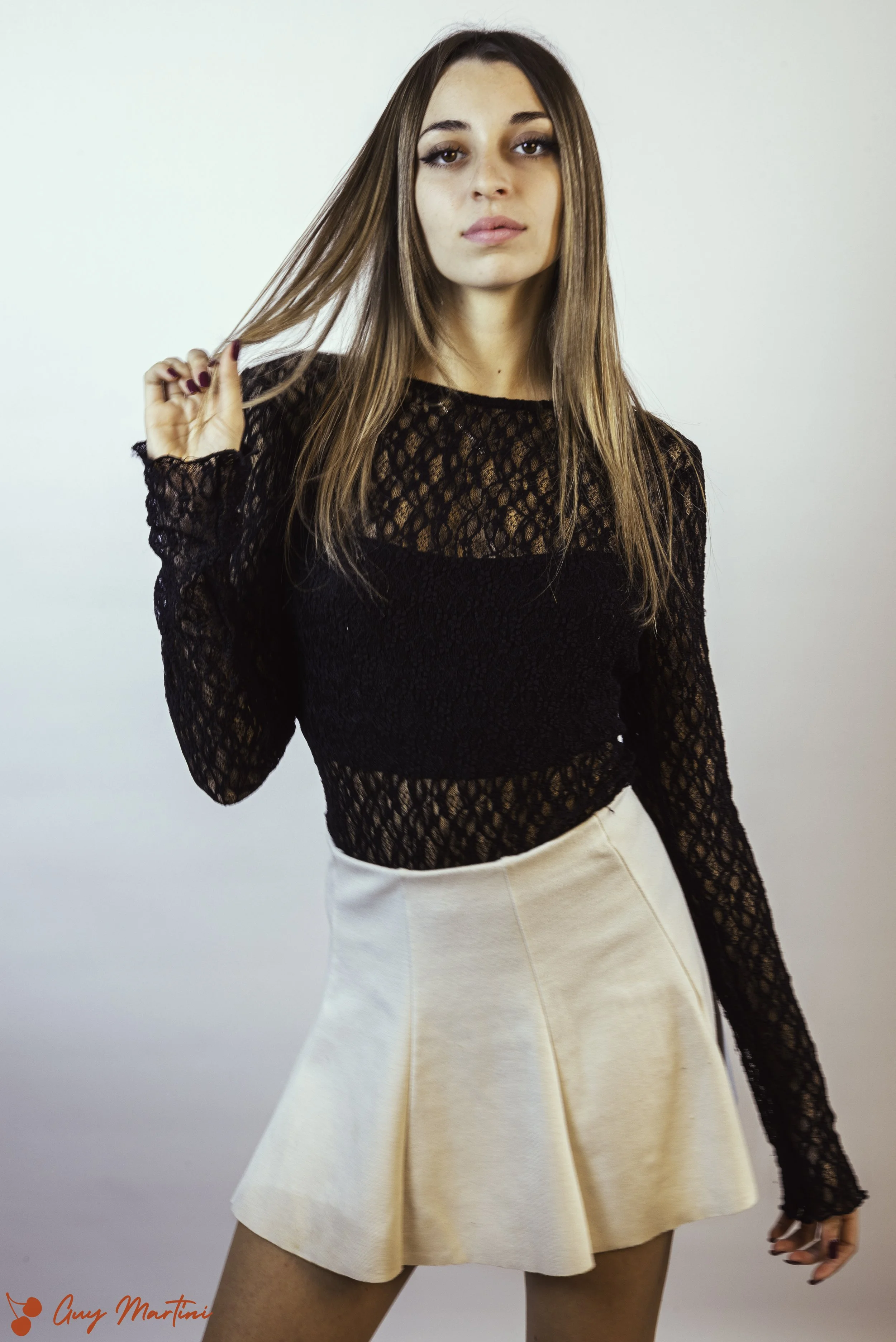 Una donna con capelli castani, occhi marroni e abbigliamento elegante, che indossa un top nero in pizzo a maniche lunghe e una gonna corta beige.