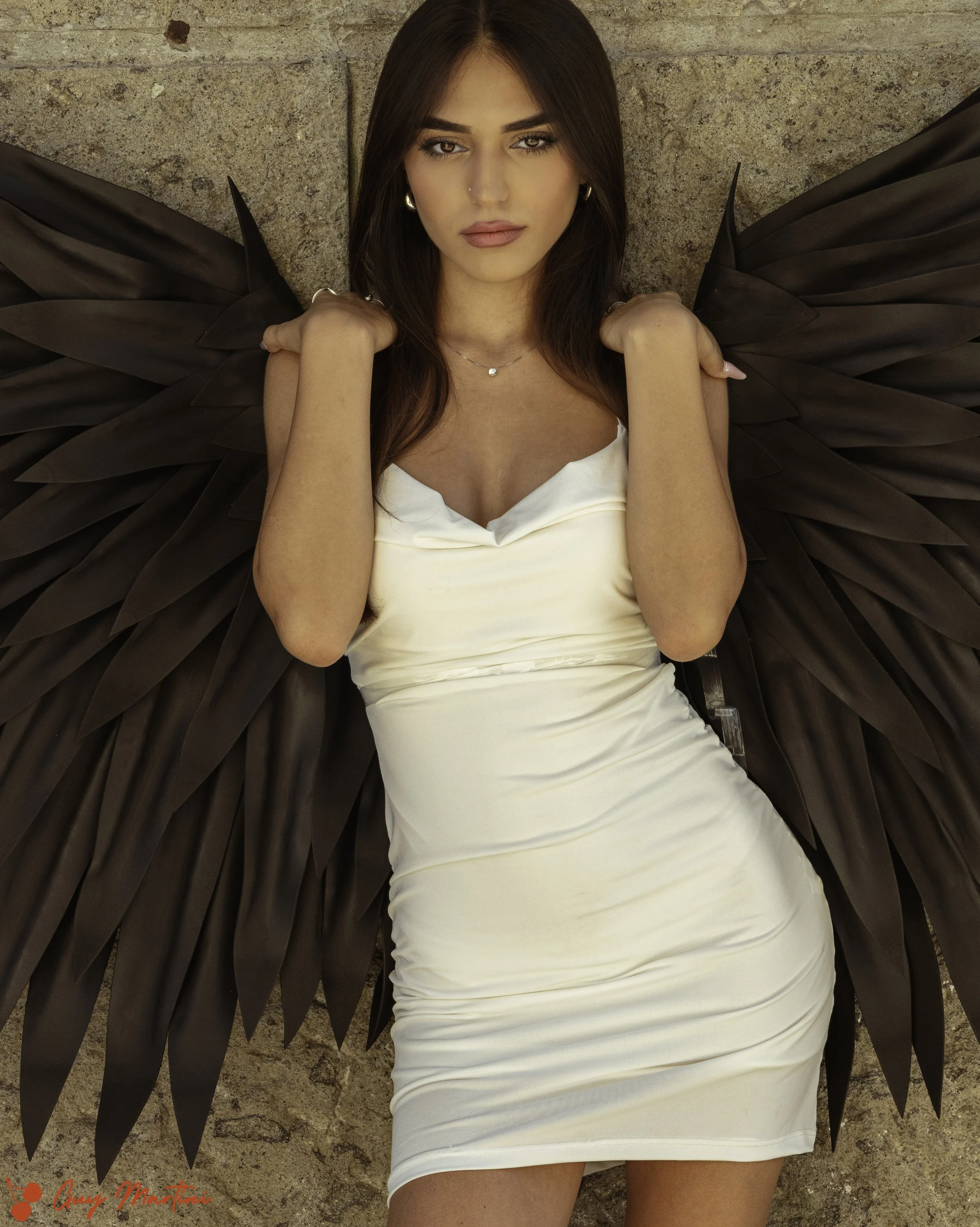 Donna con vestito bianco e grandi ali nere in posa contro un muro di pietra.