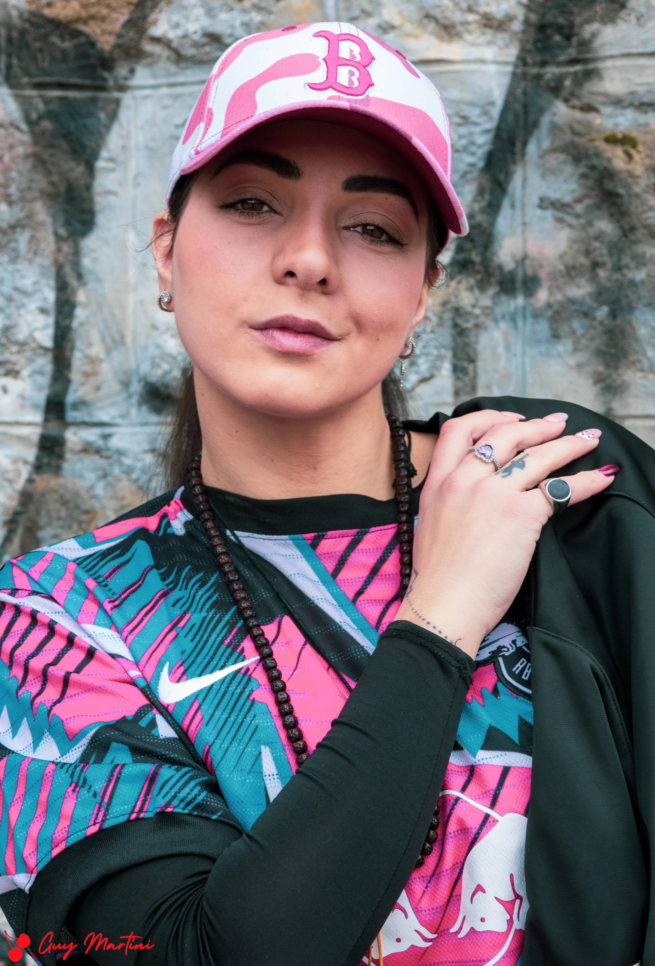 Ragazza con cappellino rosa e caschi sul capo, abbigliamento sportivo colorato, sfondo roccioso, occhialiolini argento, anelli alle dita, sorriso leggero.