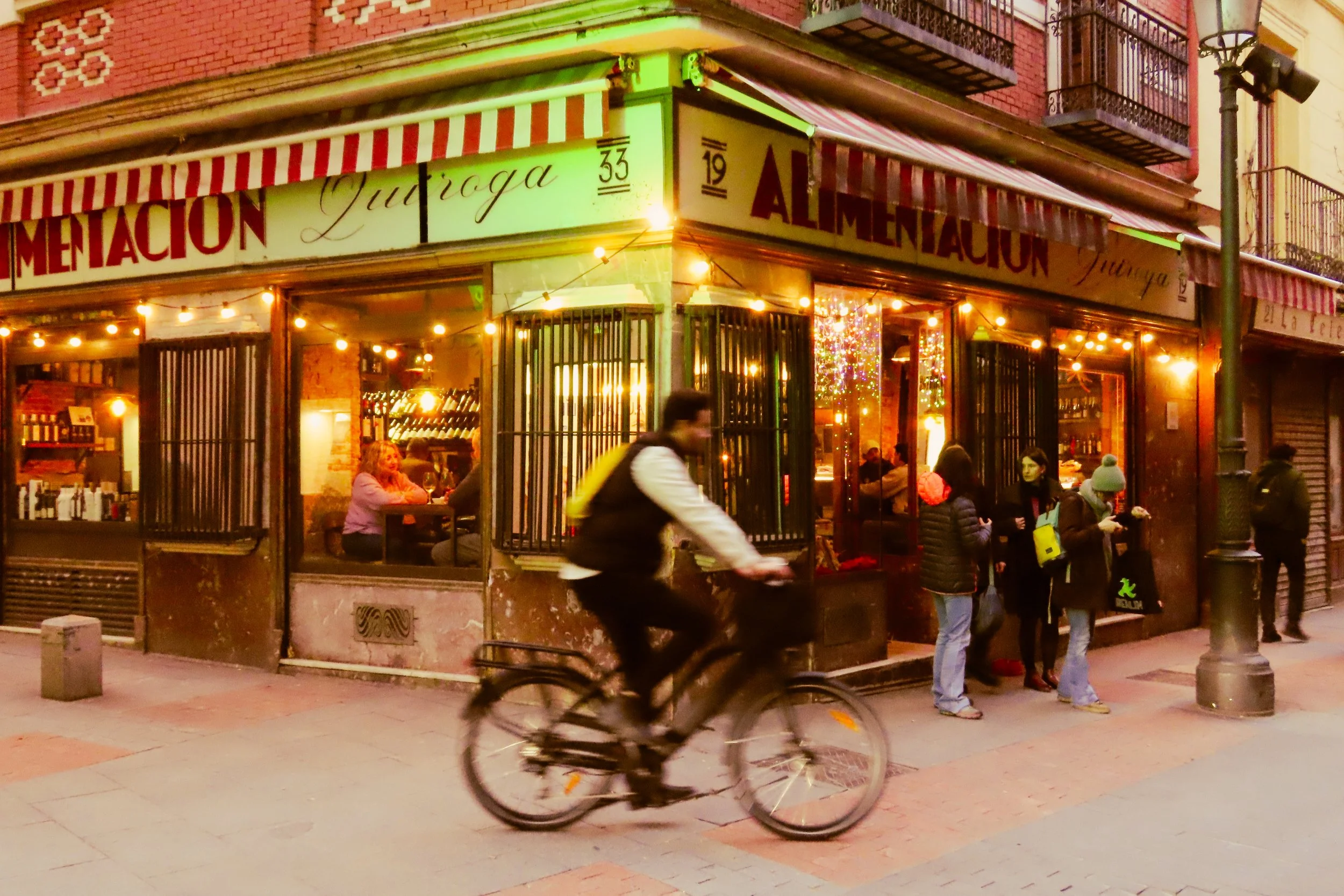 Scene di un ristorante con clienti dentro e fuori, illuminato da luci calde, con una persona che passa in bicicletta davanti, in un ambientazione urbana serale.