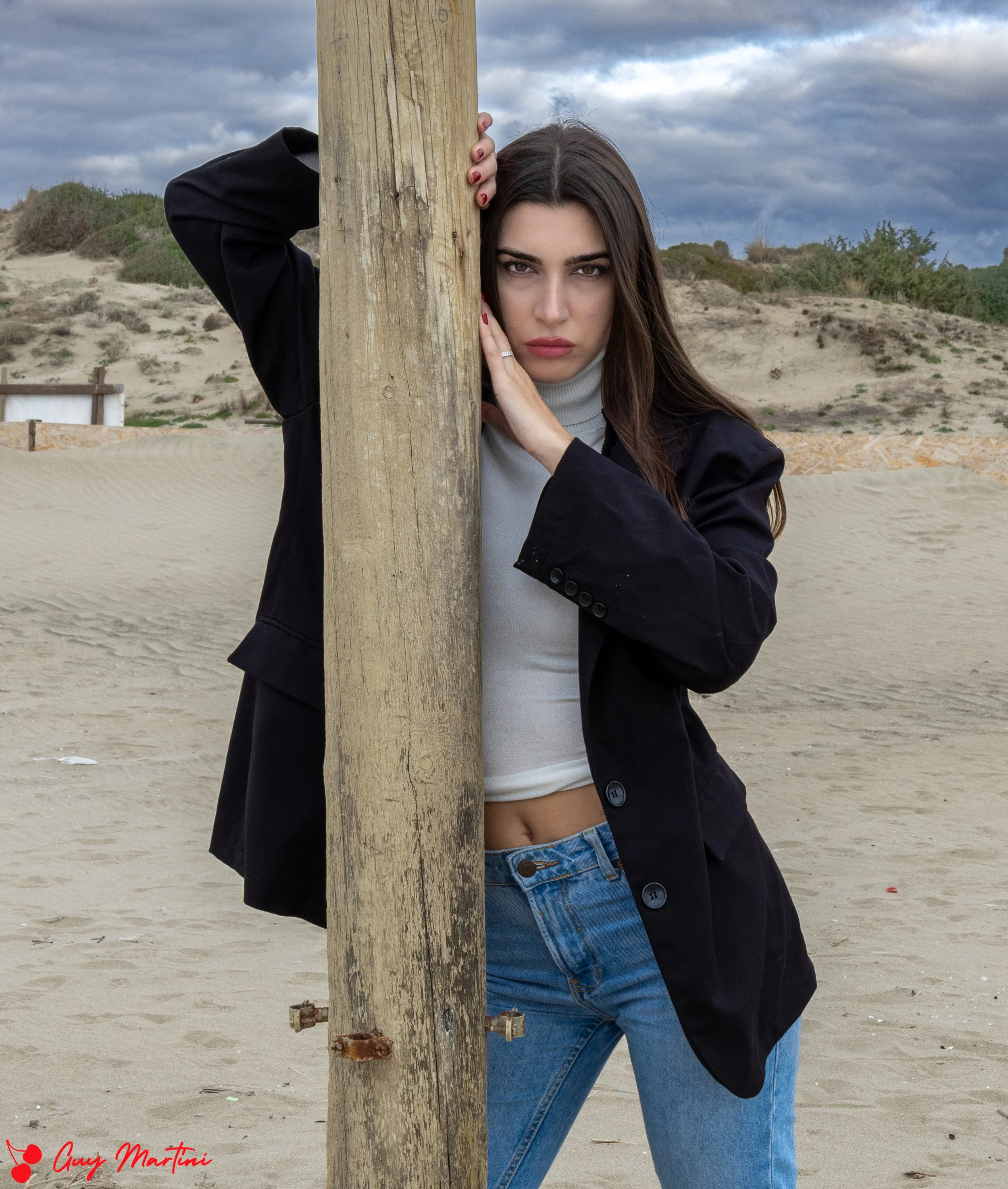 Ragazza con capelli lunghi, auricolare e vestito casual si appoggia a un palo di legno sulla spiaggia, con dune e cielo nuvoloso sullo sfondo.