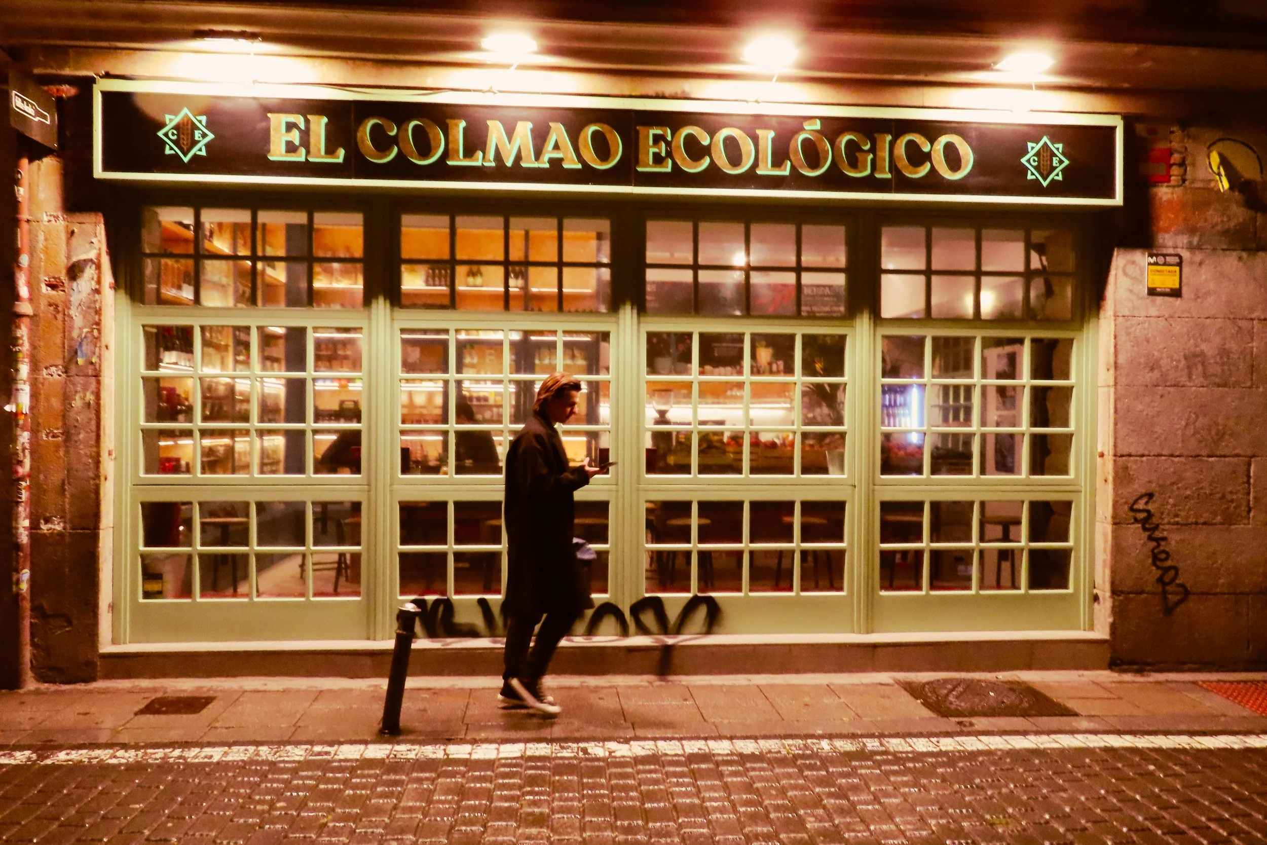 Esterno di un ristorante chiamato 'El Colmado Ecológico' con una persona che guarda il telefono in strada di sera, illuminato da luci calde, con vetrina con scaffali visibili all'interno.