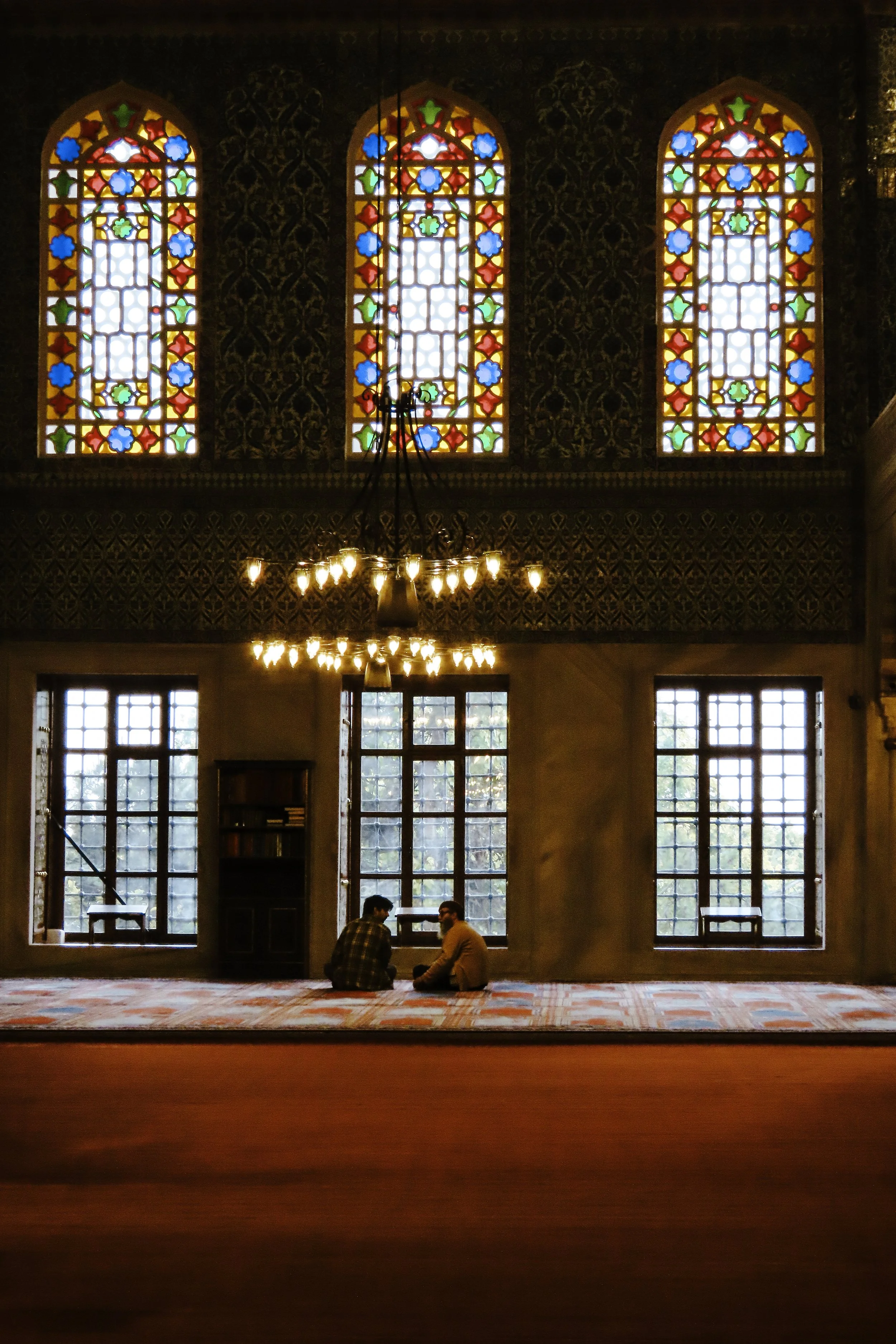 Interno di una moschea con grandi vetrate colorate in alto, lampadario e due persone sedute sul tappeto.
