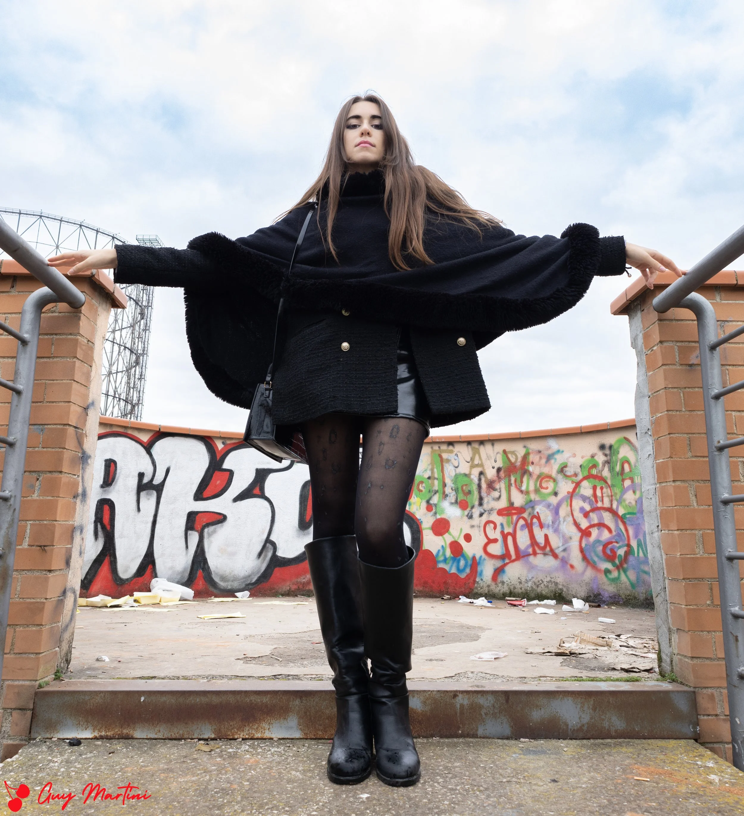 Una ragazza con un vestito nero, calze a rete con disegni e stivali neri, si trova su un ponte con graffi e murales alle pareti, con le braccia aperte