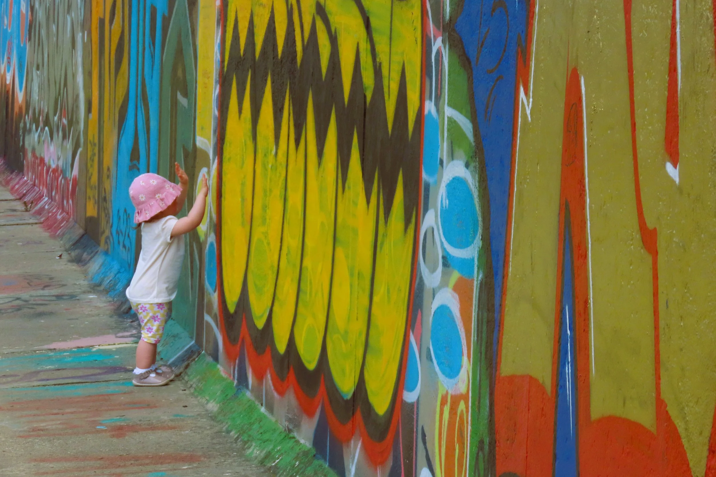 Un bambino piccolo di fronte a un murale colorato e astratto, indossa cappello rosa, maglietta bianca e pantaloncini con motivo floreale, mentre tocca o osserva la parete decorata con graffiti artistici.