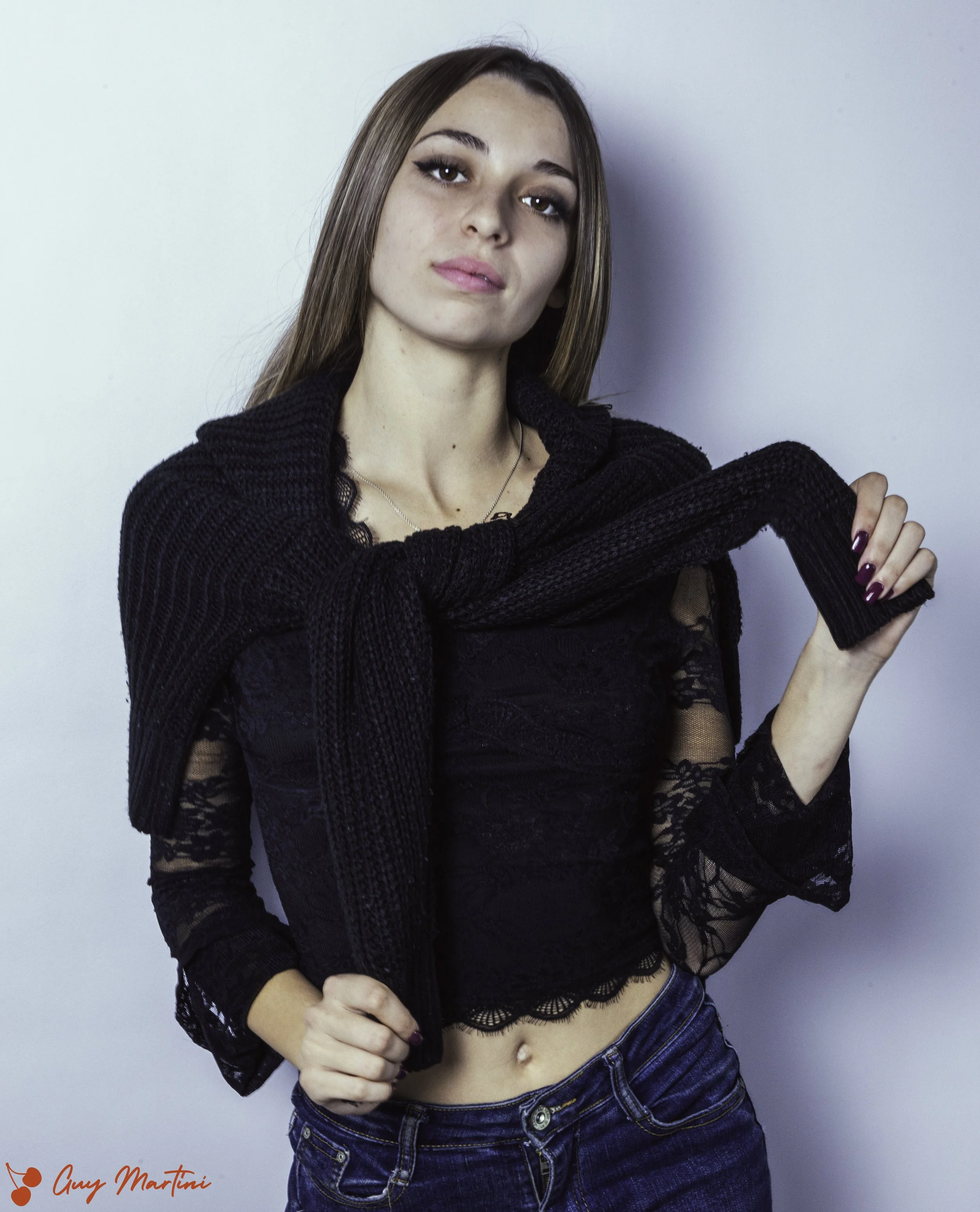 Una giovane donna con capelli castani lunghi, indossa una maglietta nera in pizzo e una felpa nera avvolta intorno alle spalle, posa davanti a uno sfondo bianco.