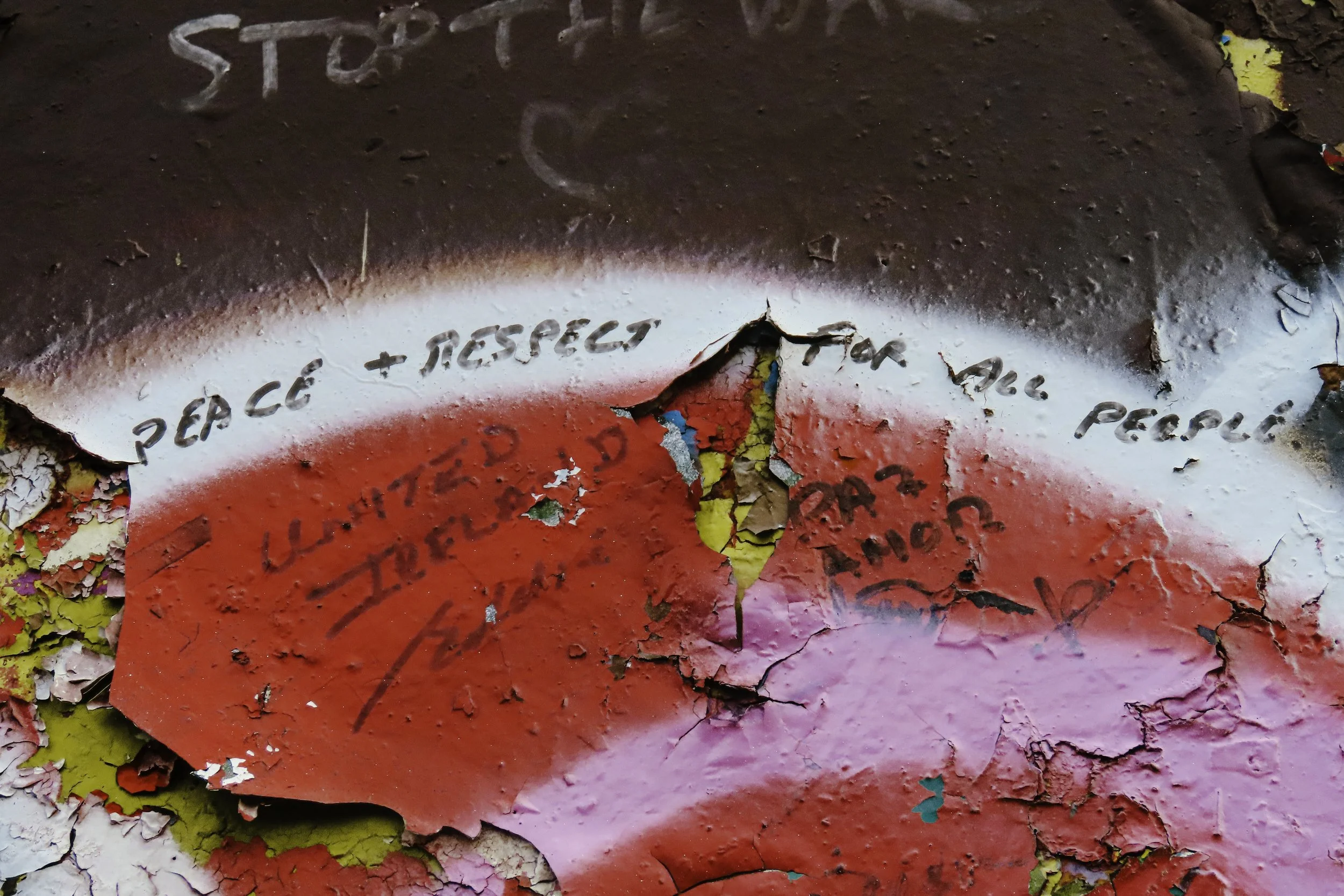 Muro con vernice spray rovinata e scritte in italiano che dicono 'Peace + Respect for all people' e altre parole non completamente leggibili, tra colori rosa, giallo e bianco con segni di crepe e peeling.